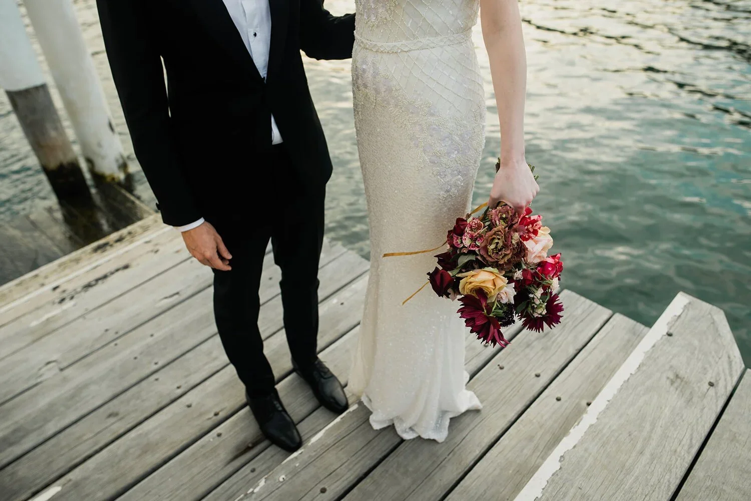 Partial view of a bride and groom standing on a wooden dock near the water, with a woman holding a bouquet of colorful flowers visible.