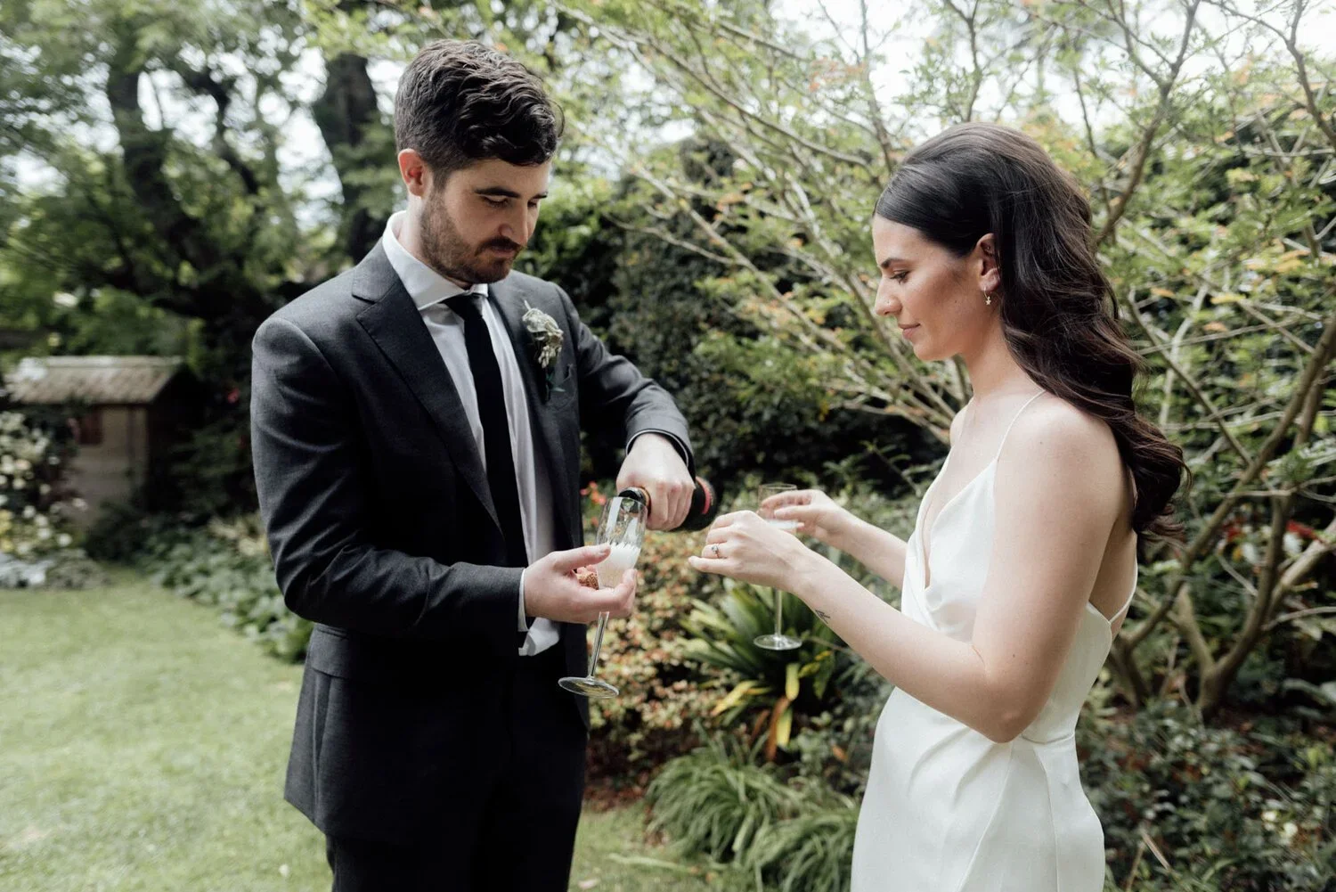 A man and woman outdoors at a wedding or celebration, the man in a gray suit and tie pouring champagne into the woman's glass, who is wearing a white dress, surrounded by greenery.