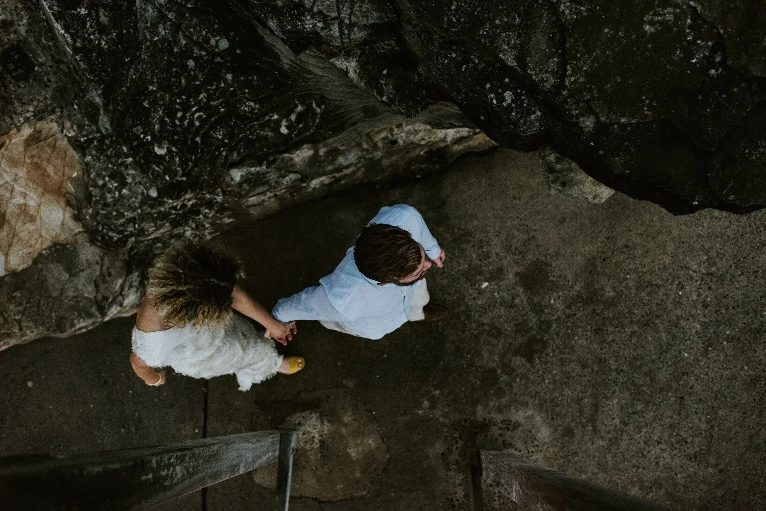 A man and a woman holding hands, seen from above, walking on a dirt path next to a large rock formation.
