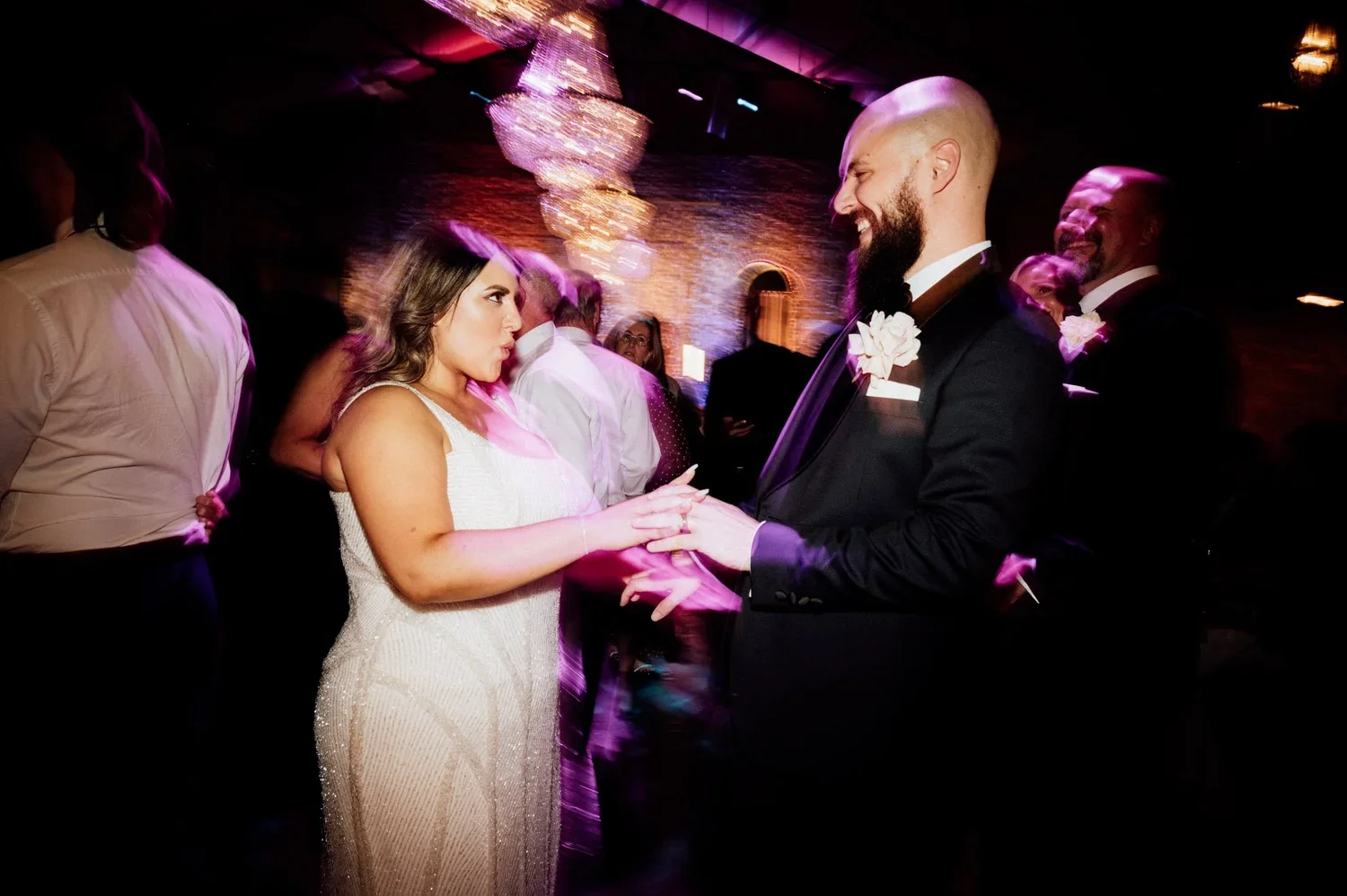 A bride and groom holding hands and dancing at their wedding reception, surrounded by guests in a dimly lit venue with purple lighting and chandeliers.