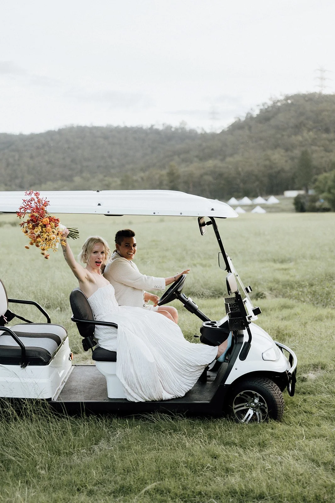 Two brides in wedding dresses sitting in a golf cart in a grassy field, one holding a bouquet of flowers and waving, with hills and tents in the background.