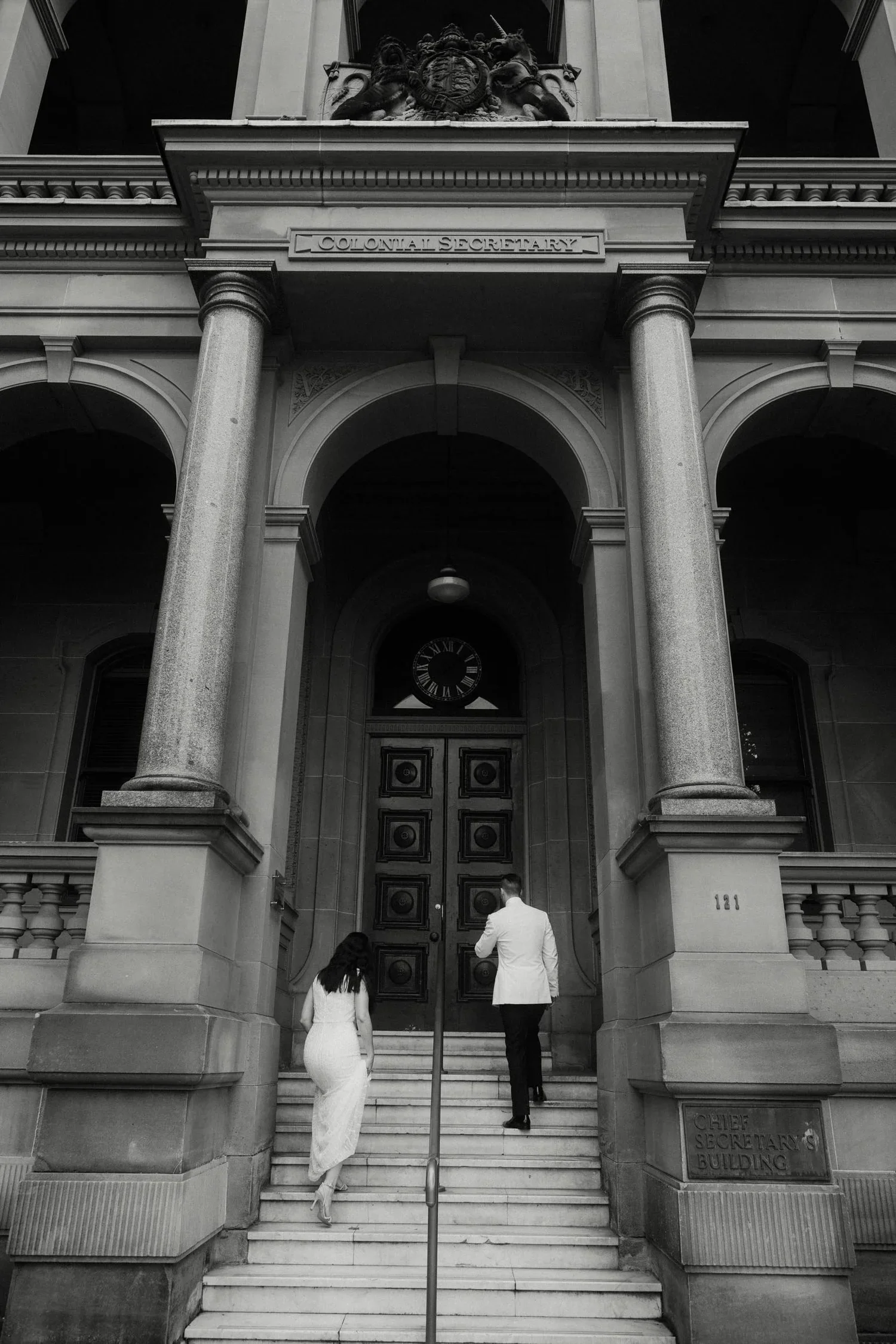 Black and white photo of a government building entrance with steps, columns, and a clock. Two people, a woman in a dress and a man in a suit, are walking up the stairs. The building has a sign that says 'Chief Secretary's Building' and another sign t