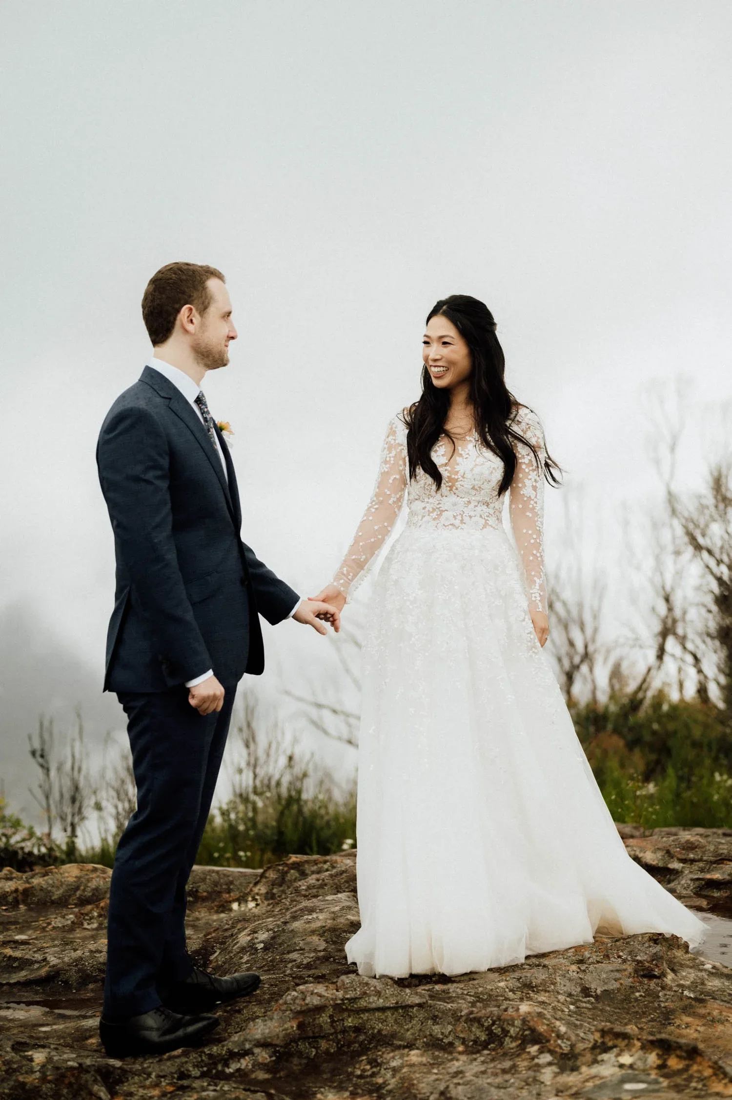A man in a dark suit and a woman in a white wedding dress holding hands outdoors on rocky ground with bare trees in the background.