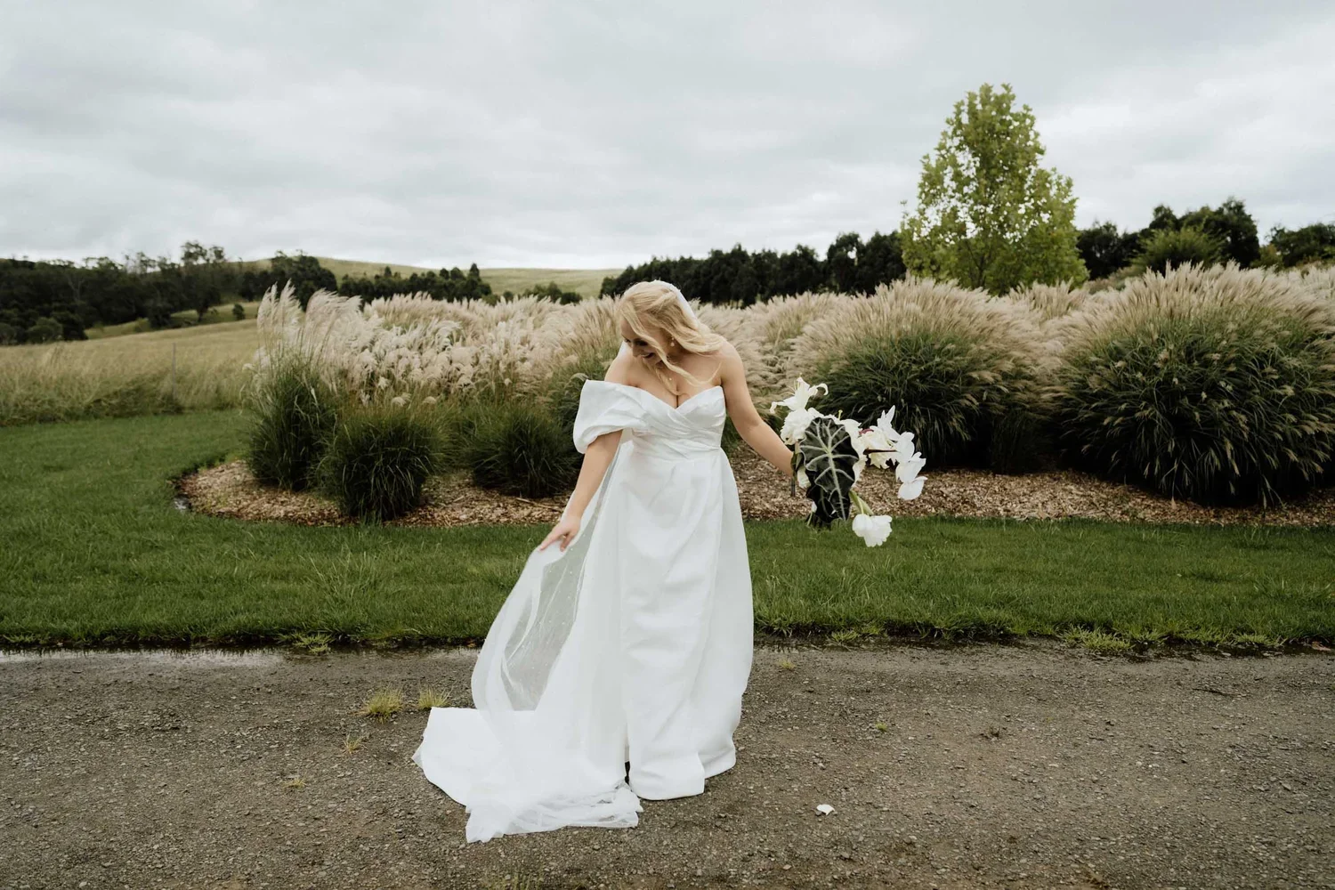A woman in a white wedding dress holding a bouquet of white flowers, standing outdoors on a gravel pathway with lush green grass, tall grasses, and trees in the background on a cloudy day.