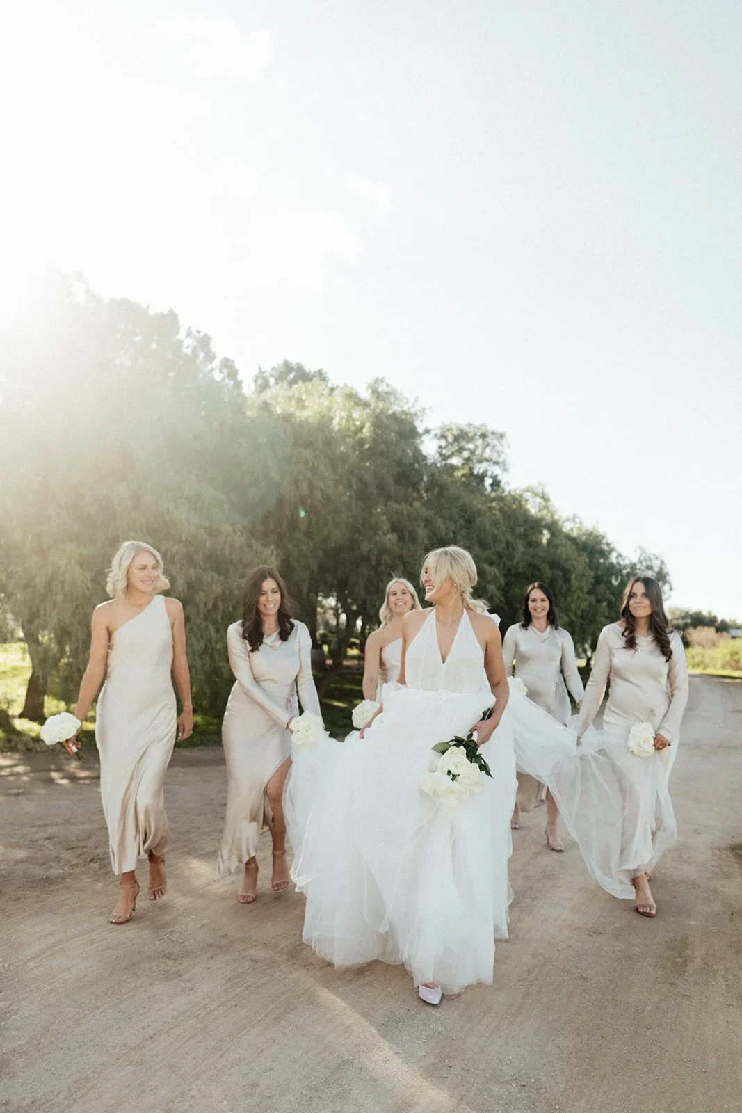 A group of six women walking outdoors on a sunny day, all dressed in white with some holding flowers, in a natural setting with trees in the background