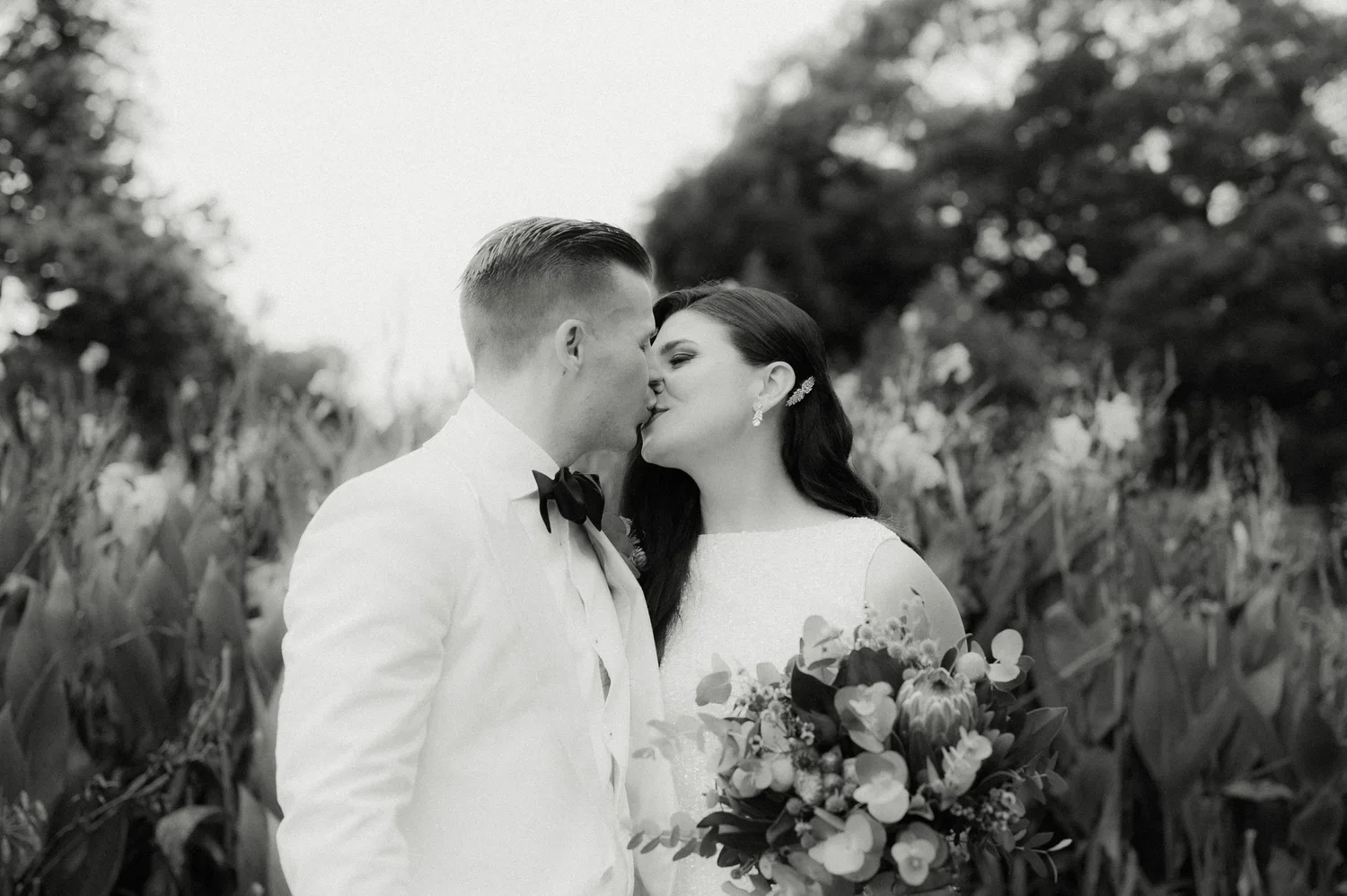 A black-and-white photo of a bride and groom kissing outdoors, with the bride holding a bouquet of flowers, in a field of tall plants, with trees in the background - Sydney Botanical gardens wedding photography.