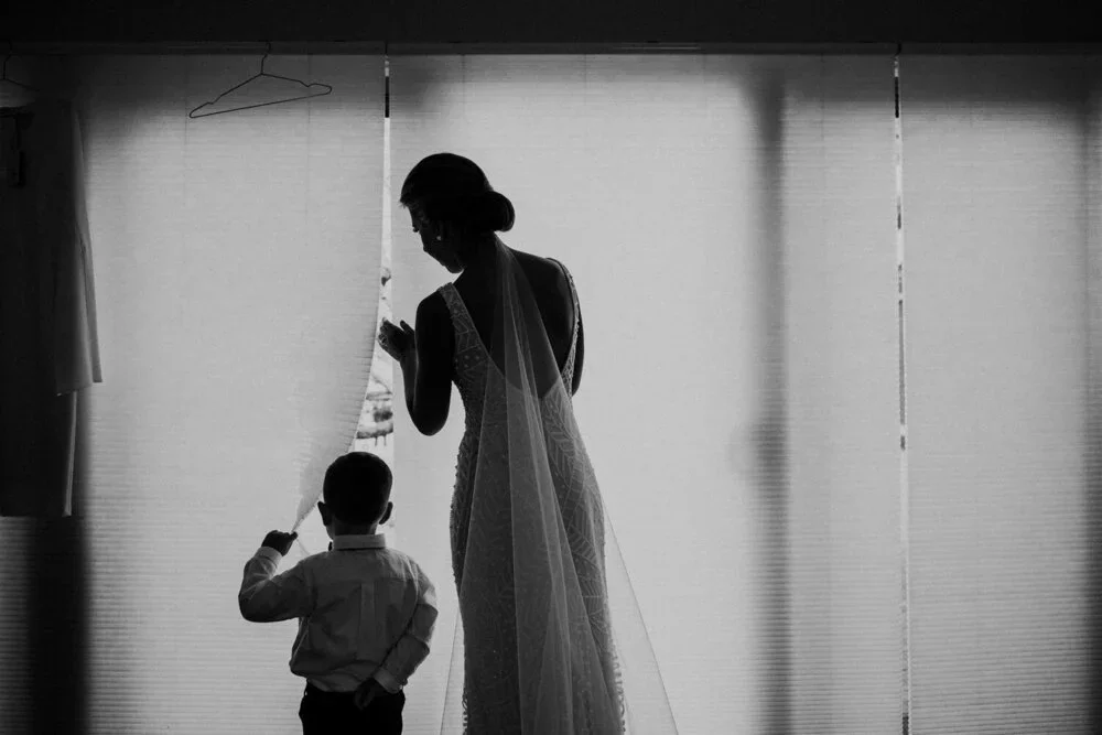 A woman in a wedding dress and veil stands behind a young boy, both looking out a partially open curtain.