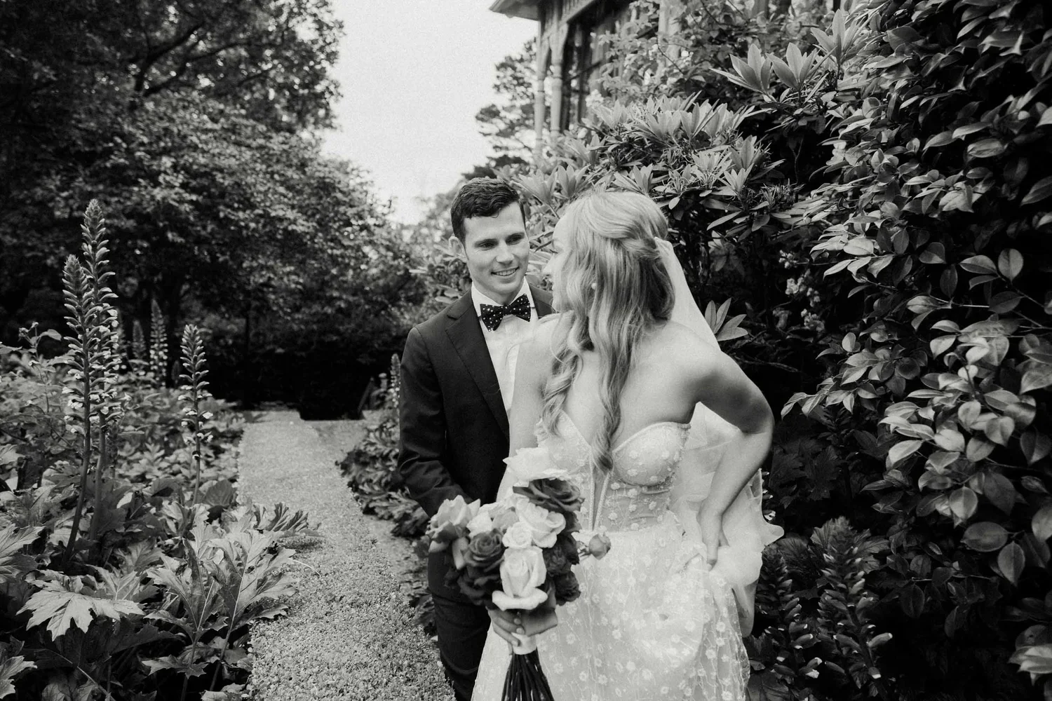 A bride and groom standing together outdoors, smiling at each other, with the bride holding a bouquet of roses and wearing a strapless lace wedding dress, surrounded by lush green plants and trees.