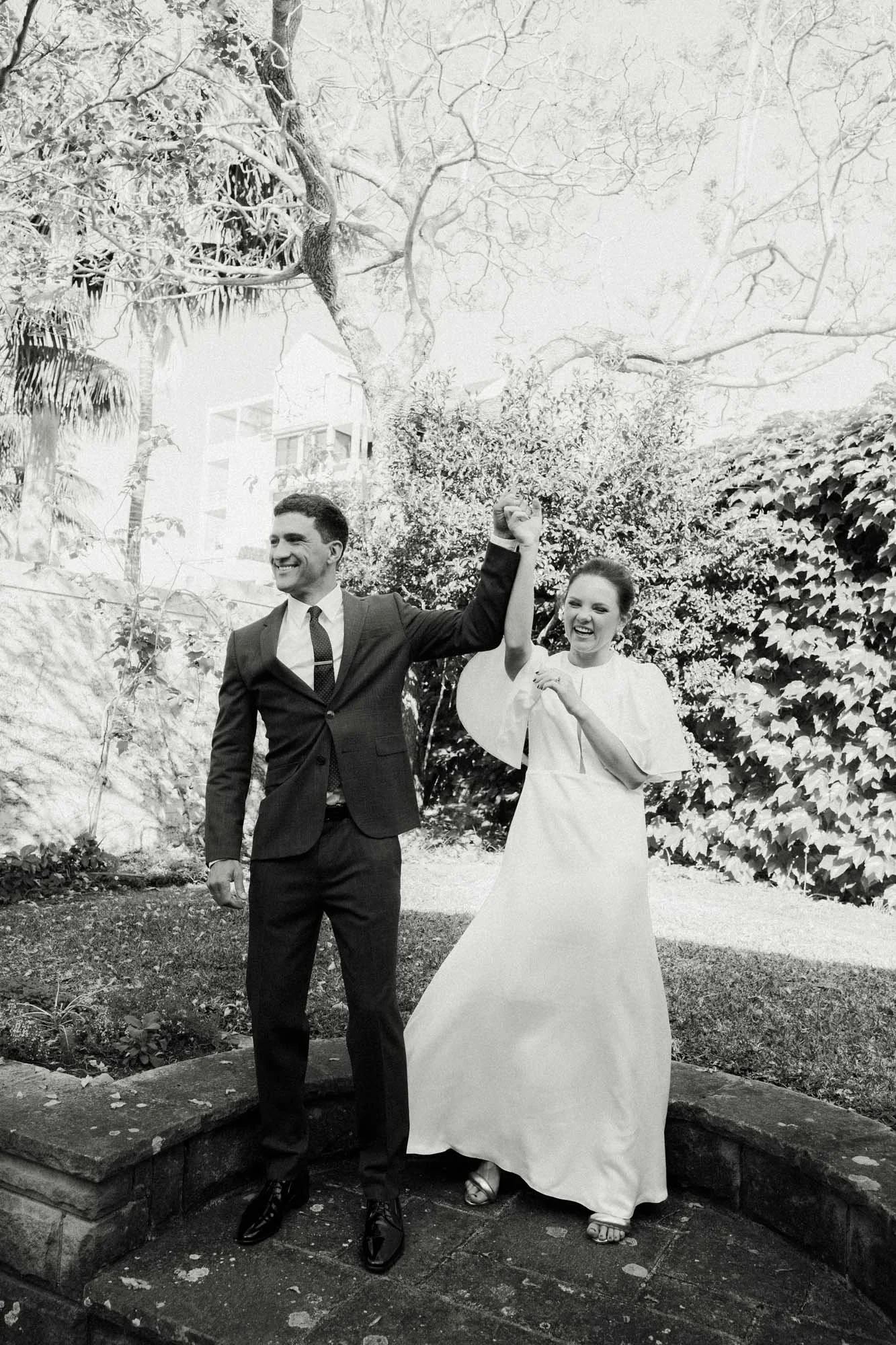 A black and white photo of a smiling couple walking outdoors, holding hands, with trees and bushes in the background.