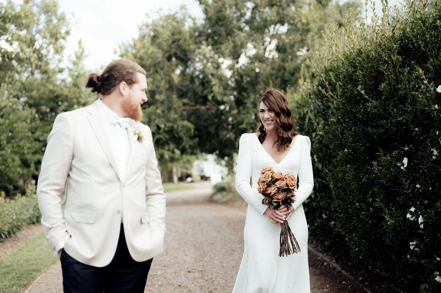 A bride with brown hair in a white wedding gown holding a bouquet of brown roses, smiling at her groom, a man with long hair and a beard in a white suit, on a garden path surrounded by green trees and bushes.