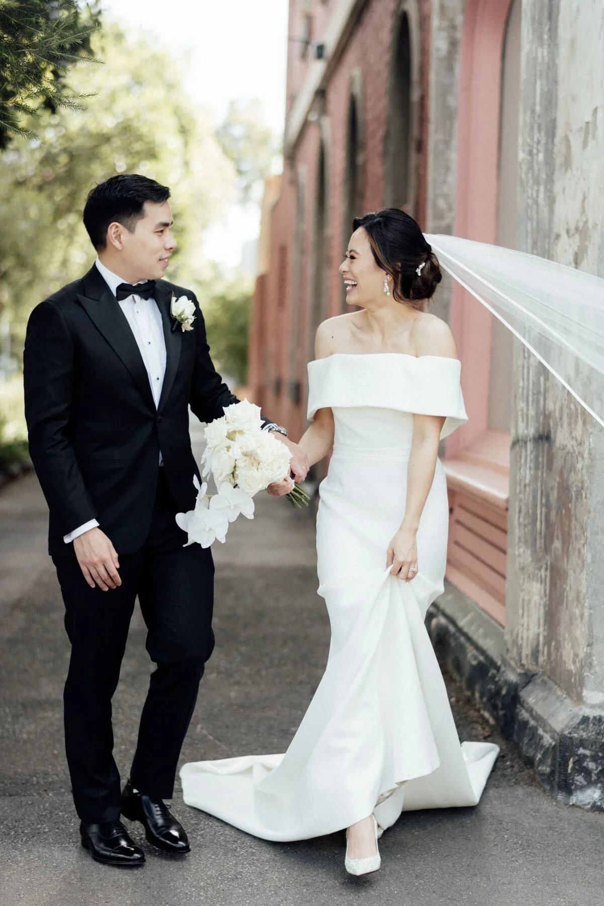 A bride and groom on a city street, holding hands and smiling at each other. The bride wears a white off-the-shoulder wedding gown with a long train and veil, and the groom wears a black tuxedo with a bowtie. The groom holds a bouquet of white flower