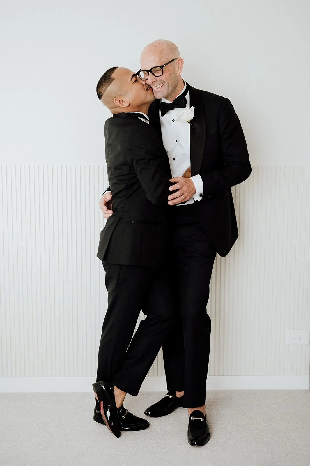 Two grooms on their wedding day in tuxedos hugging and smiling at each other, one wearing glasses and the other with short hair, against a plain white background.