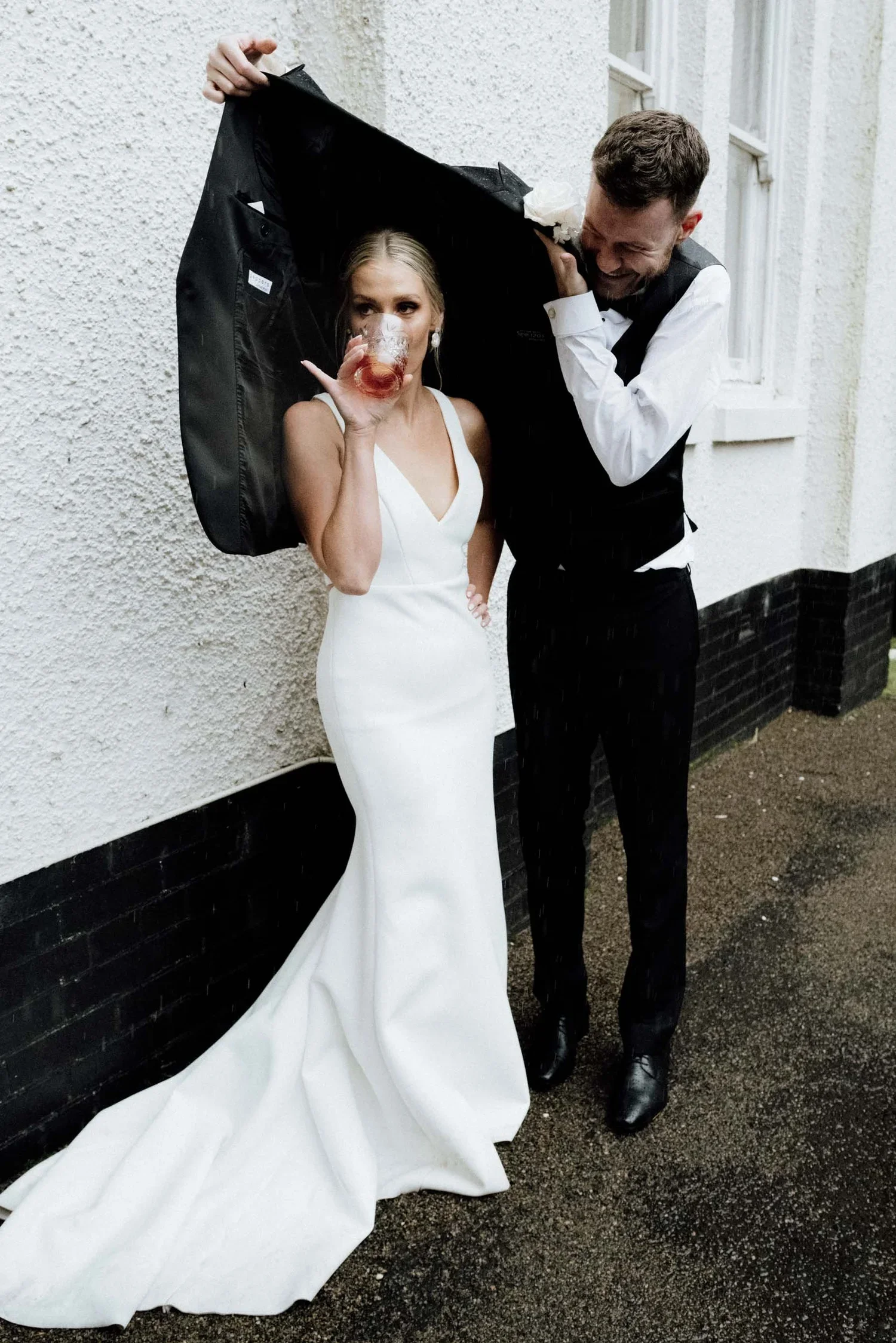 A bride in a white wedding dress with a long train drinking from a glass while a man, possibly the groom, in a black vest and white shirt laughs and holds an umbrella over her. They are standing outside against a white, textured wall with a window.