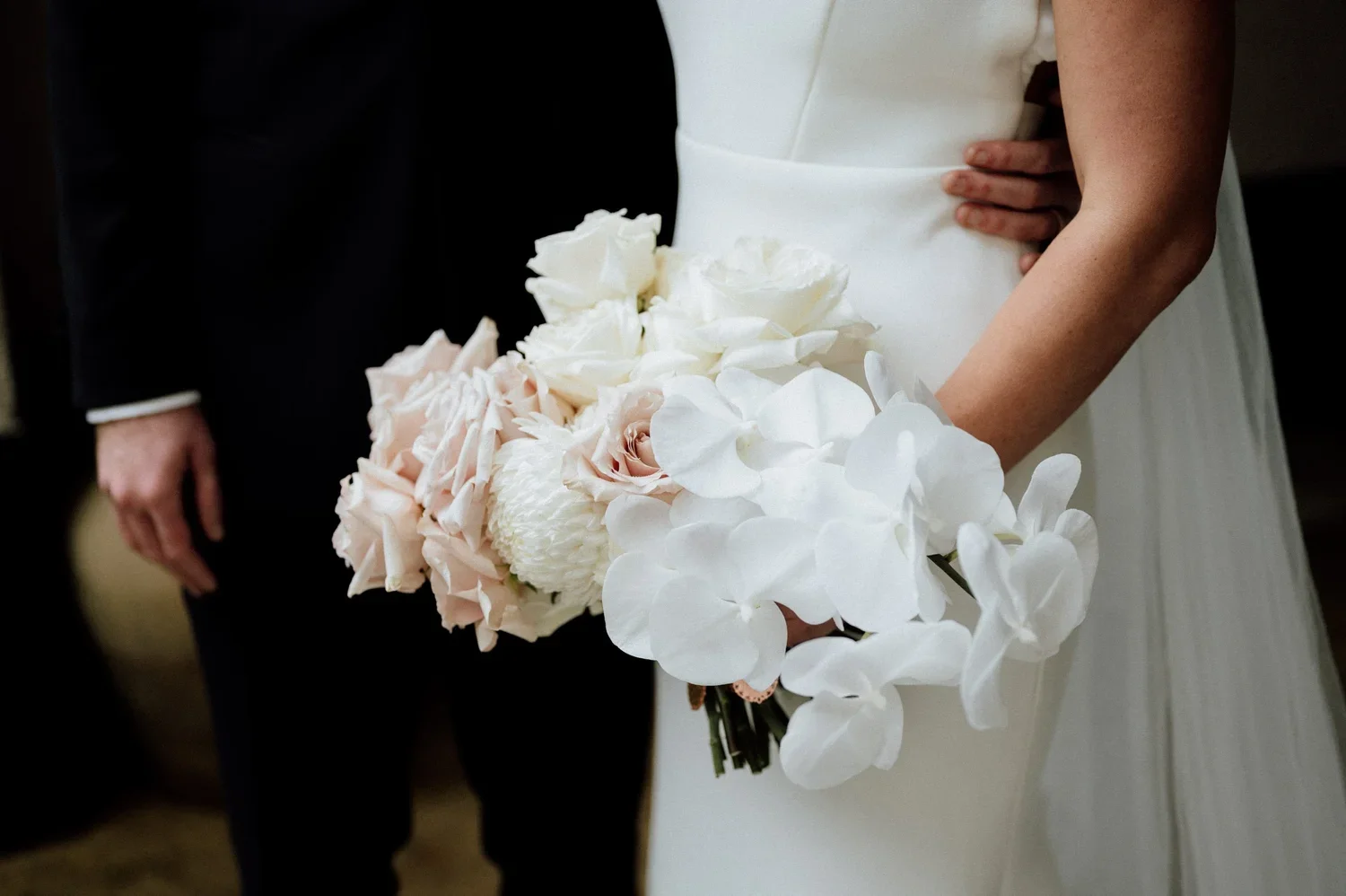 A bride holding a bouquet of white and blush pink flowers, including roses and orchids, during a wedding ceremony.