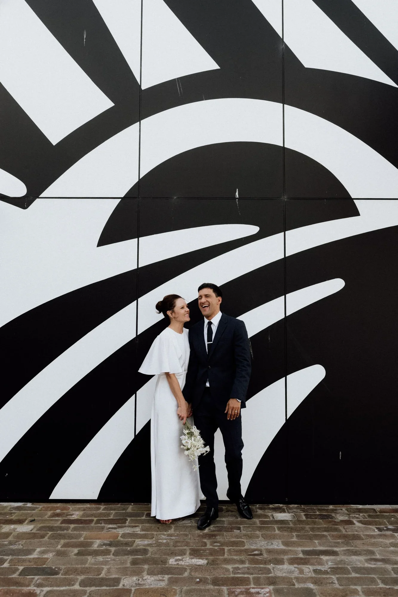 A bride and groom stand together in front of a large black-and-white mural with abstract lines and shapes. The bride wears a white dress and holds a bouquet, while the groom wears a dark suit and tie. They are smiling and looking at each other.
