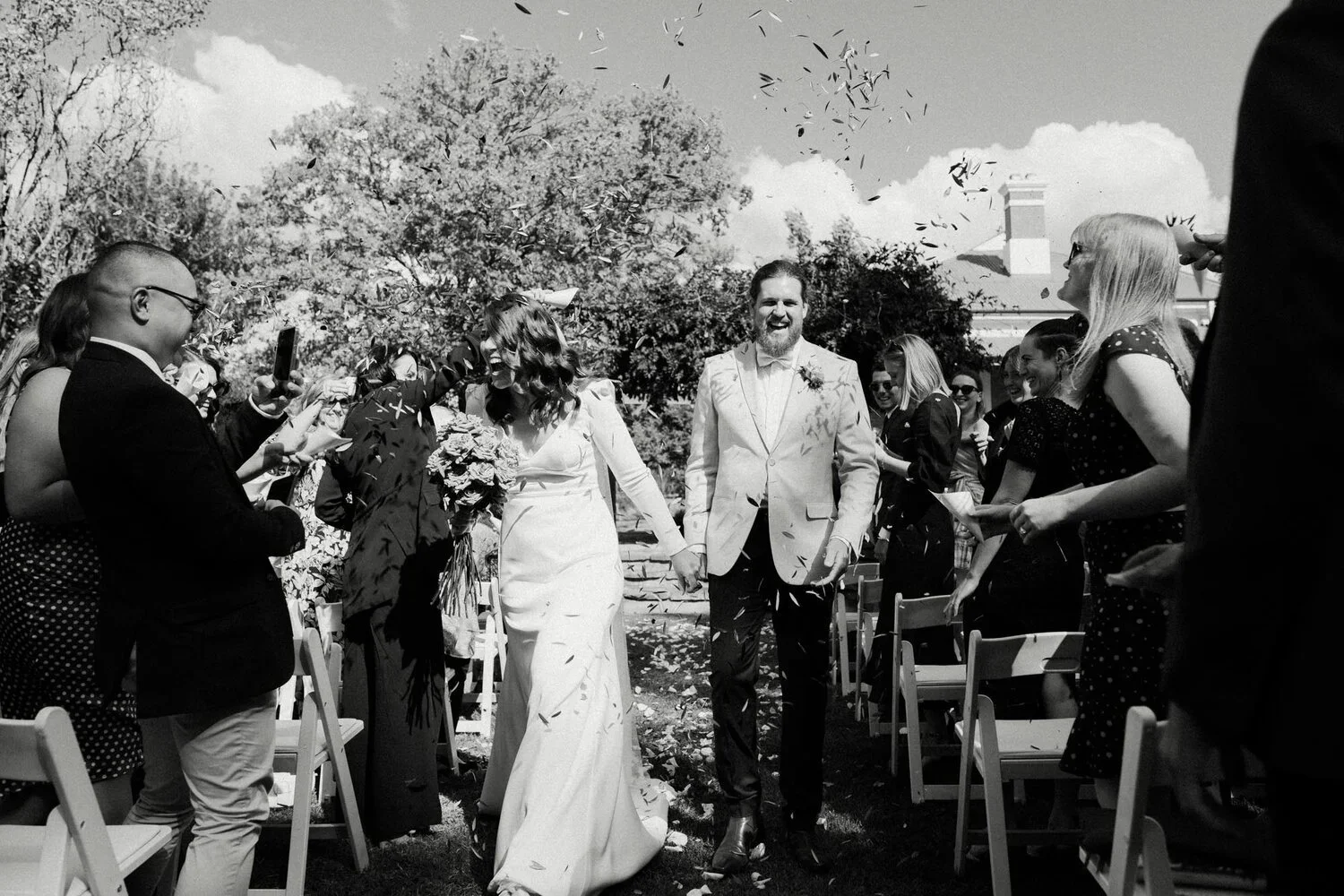 Black and white photo of a wedding ceremony outdoors, showing a bride and groom holding hands and smiling, walking down the aisle amidst a crowd of guests cheering and throwing confetti.