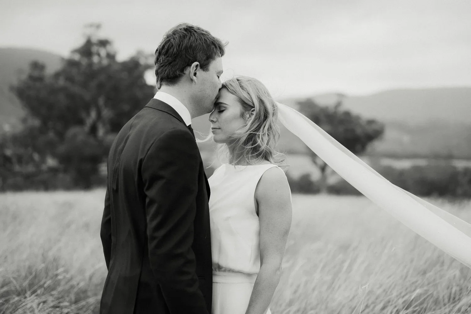 A black-and-white photo of a bride and groom standing close together in a field. The groom is kissing the bride on her forehead, and the bride has eyes closed with a serene expression. The bride wears a sleeveless dress and a long veil blowing in the