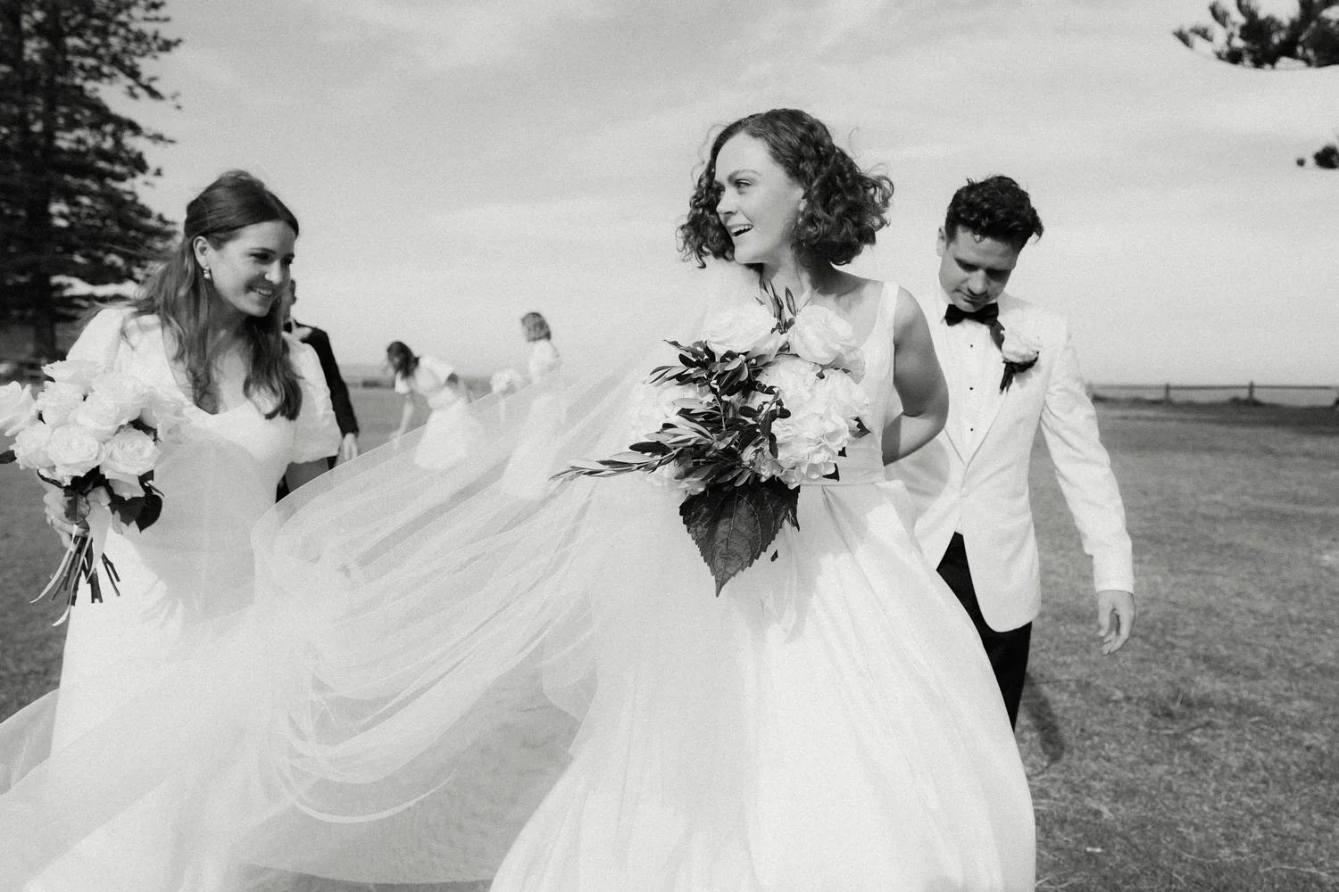 Black and white photo of a wedding party outdoors, featuring a bride with curly hair holding a bouquet of flowers, a groom in a tuxedo, and two bridesmaids in dresses holding bouquets, walking on grass with a distant horizon and trees in the backgrou