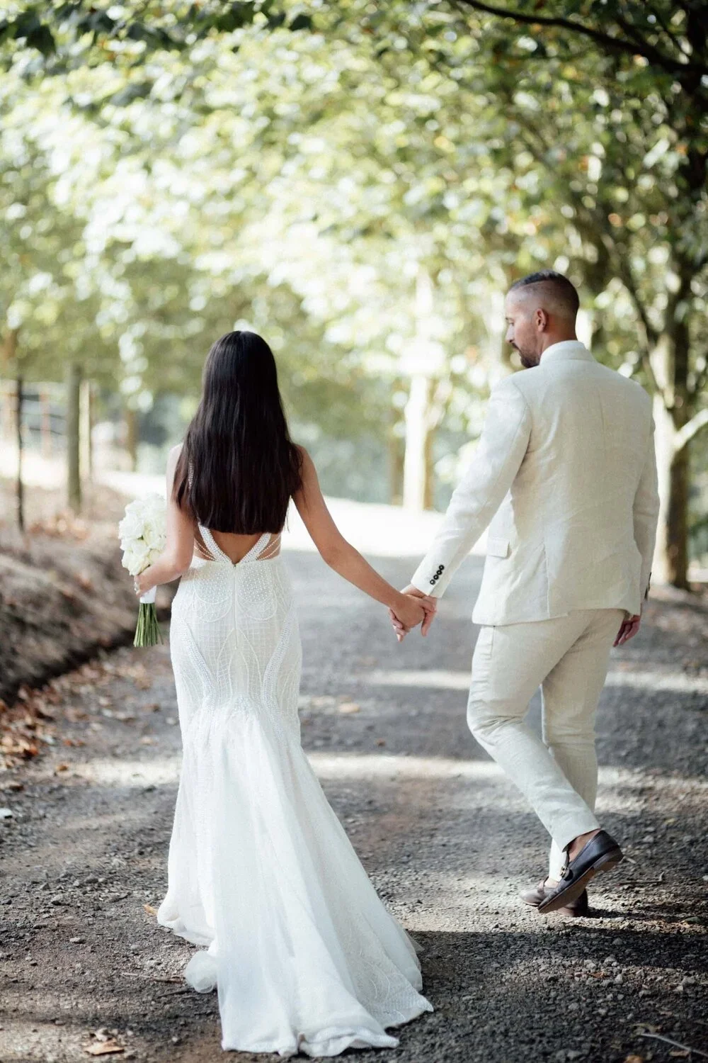 A bride and groom holding hands and walking down a tree-lined dirt path during their wedding day. The bride is wearing a white, backless wedding gown and holding a bouquet of white flowers. The groom is wearing a light-colored suit with dark shoes.