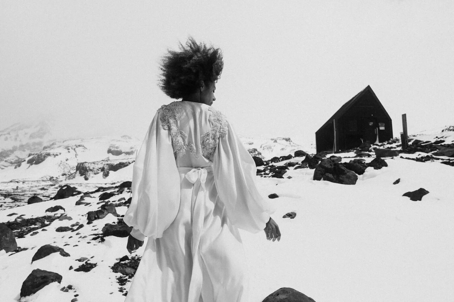 A woman in a flowing, embroidered white dress walking through a snow-covered landscape, with a small dark wooden house in the background.