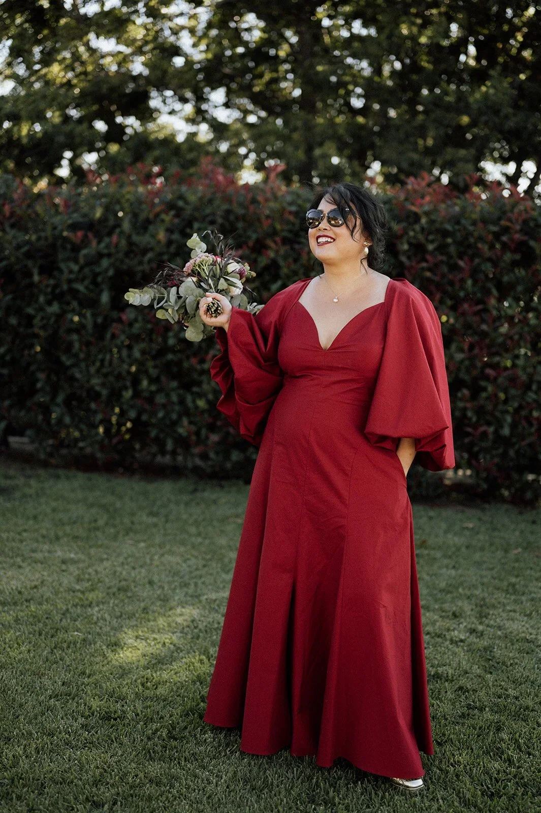 A woman in a long red dress holding a bouquet of flowers, wearing sunglasses, standing outdoors on grass with a background of bushes and trees, smiling - non traditional Chinese wedding dress and photographer in Sydney. 
