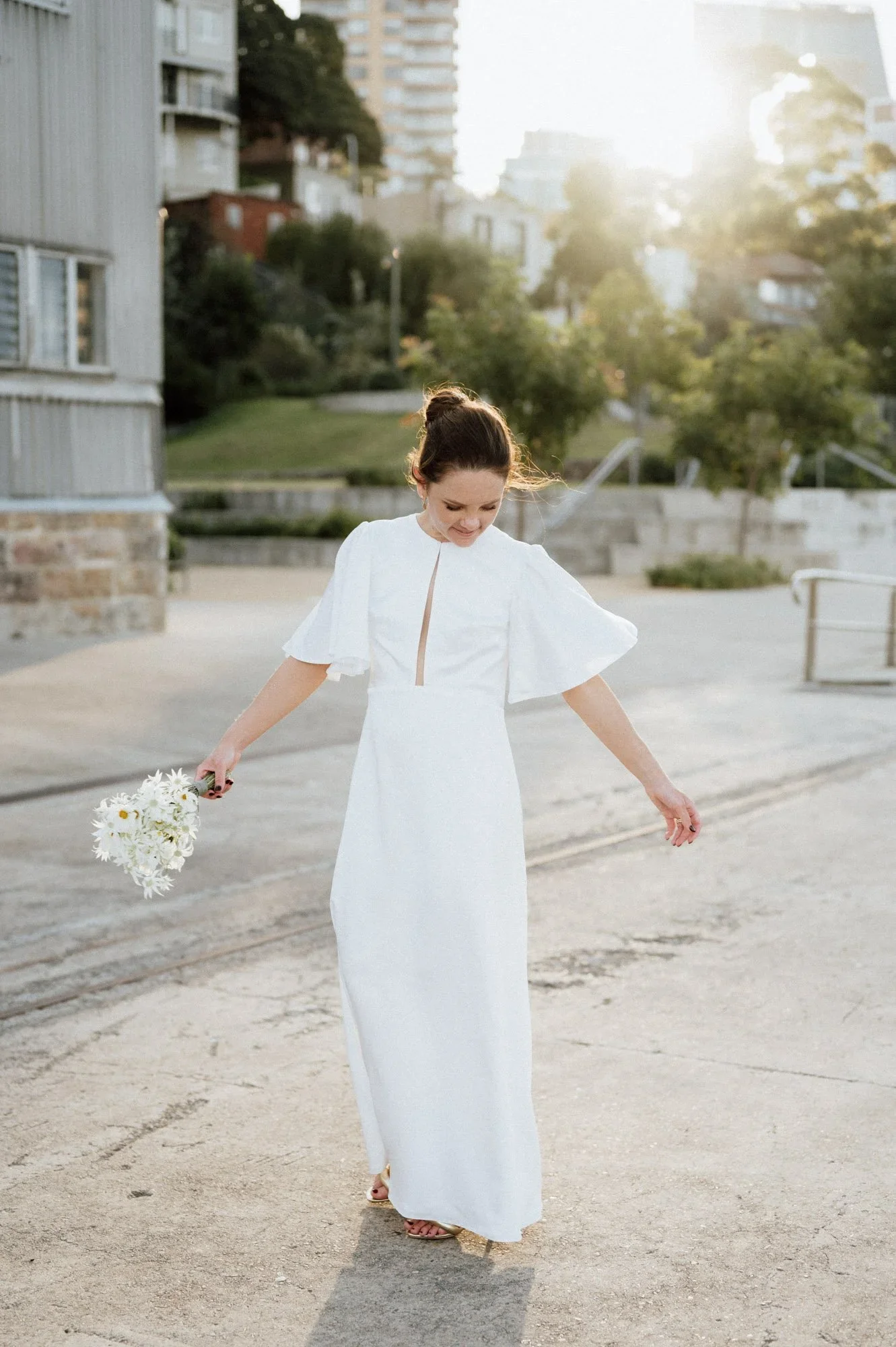 A woman in a long white dress with short sleeves stands outdoors on a paved surface, holding a bouquet of white flowers, with her head slightly bowed and smiling, sunlight shining behind her, and a background of trees and multi-story buildings.