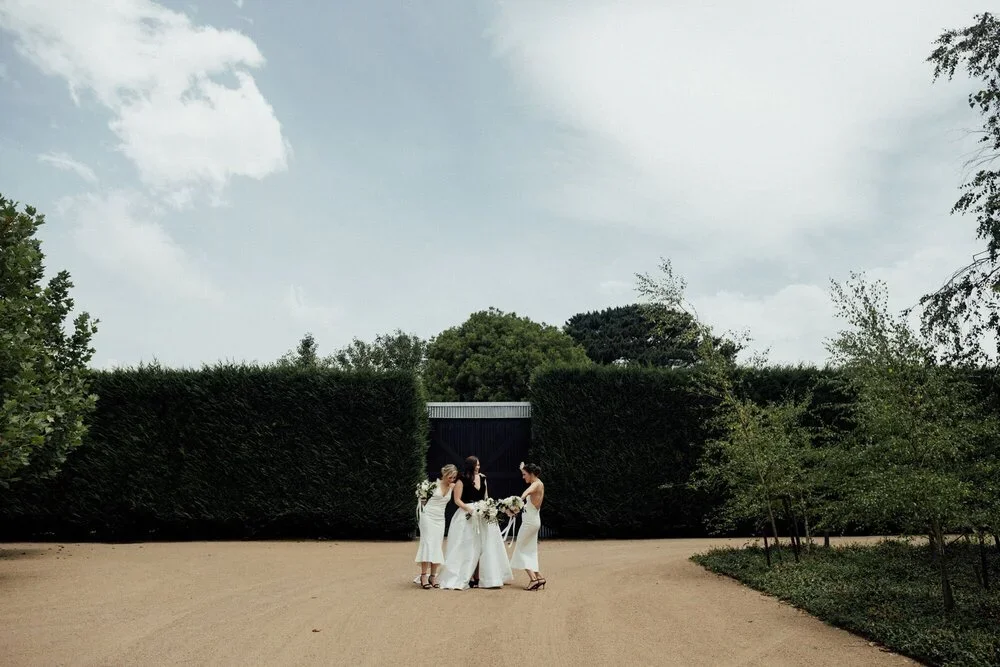 Four women, two in white wedding dresses and two in black tops and white skirts, standing on a dirt path in front of a large hedge, possibly in a garden or park setting, with trees and a cloudy sky above.