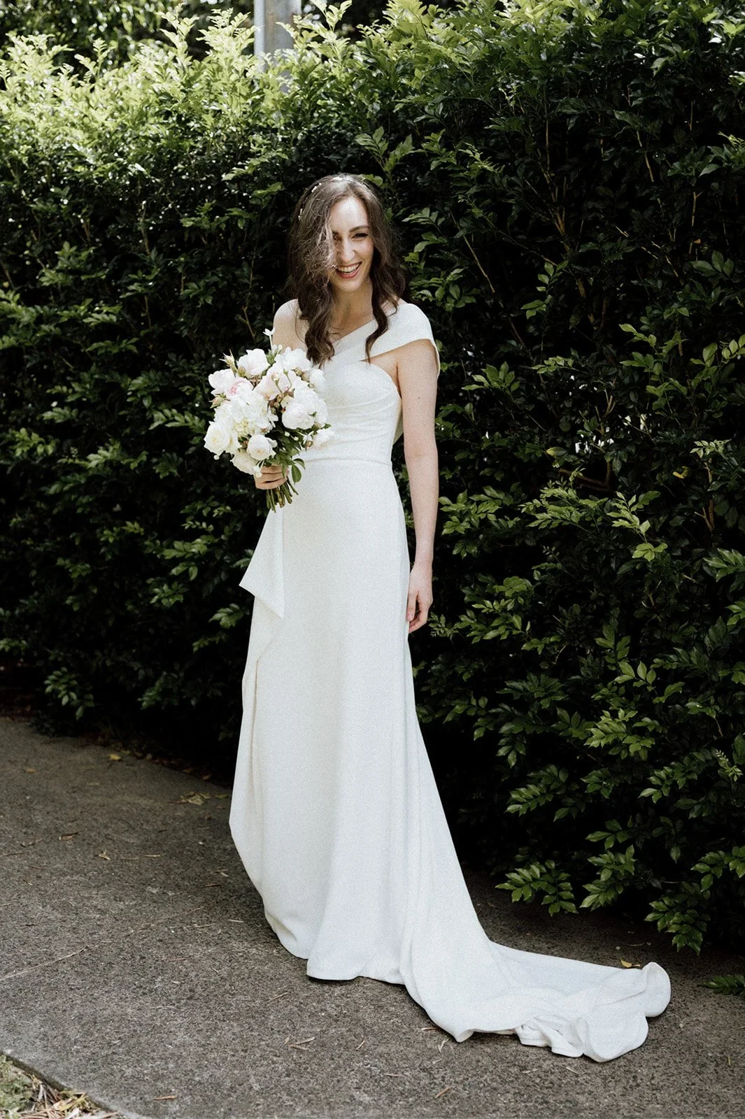 A woman wearing a white wedding dress, holding a bouquet of white flowers, standing outside on a path with green bushes in the background, smiling and looking joyful.
