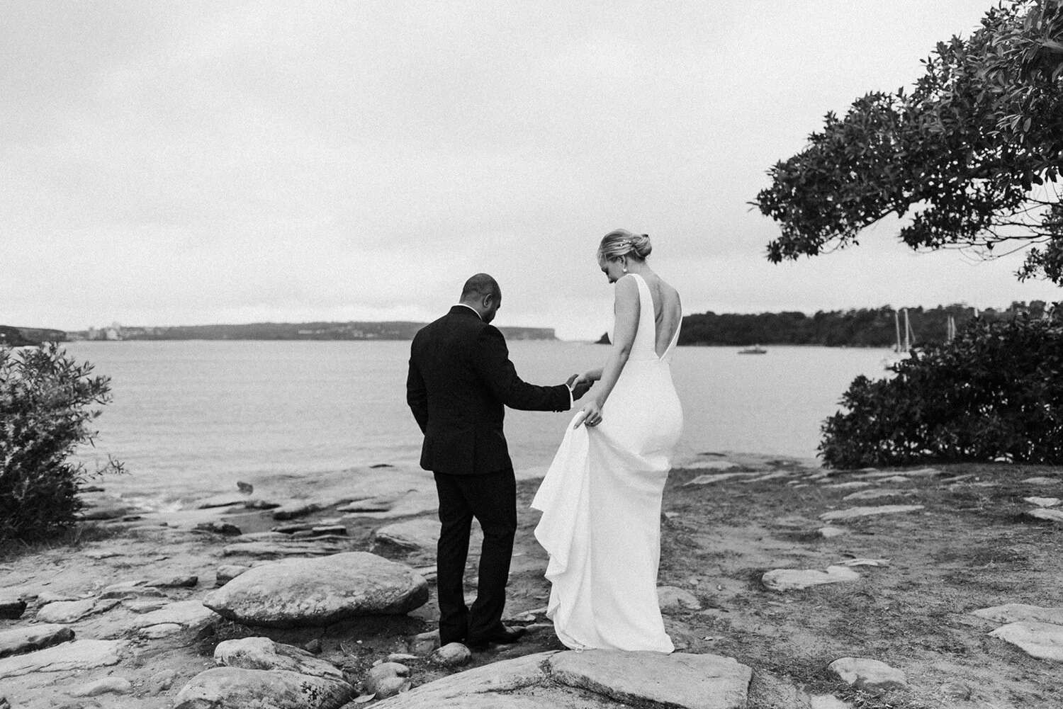 A black-and-white photo of a bride and groom holding hands on a rocky beach by the water, with trees and boats in the background.