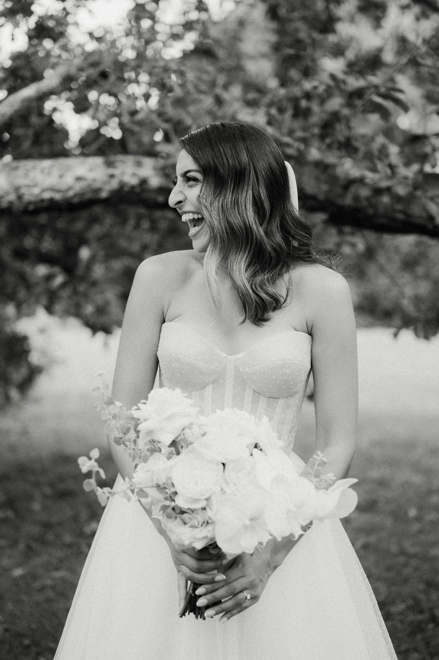 Black and white photo of a woman smiling and looking to her left, holding a bouquet of flowers, outdoors with trees in the background.