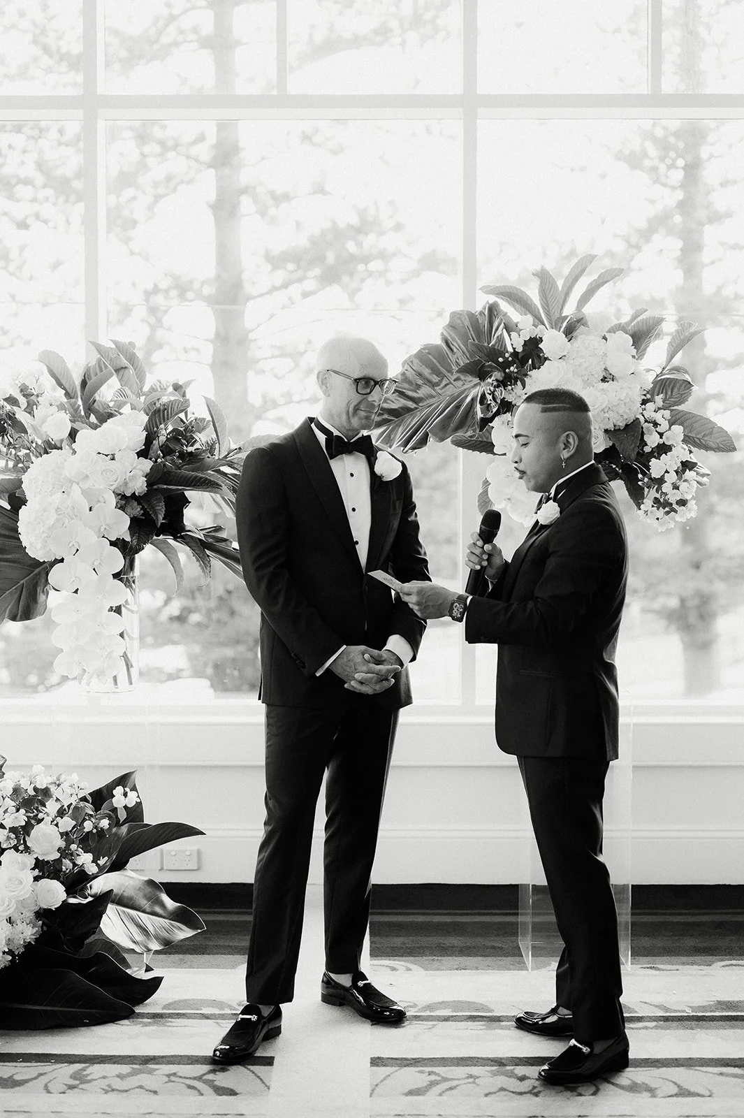 Two men in tuxedos with boutonnieres participating in a wedding ceremony. One person is reading from a piece of paper while holding a microphone. The setting includes large floral arrangements and a large window in the background, inclusive wedding.