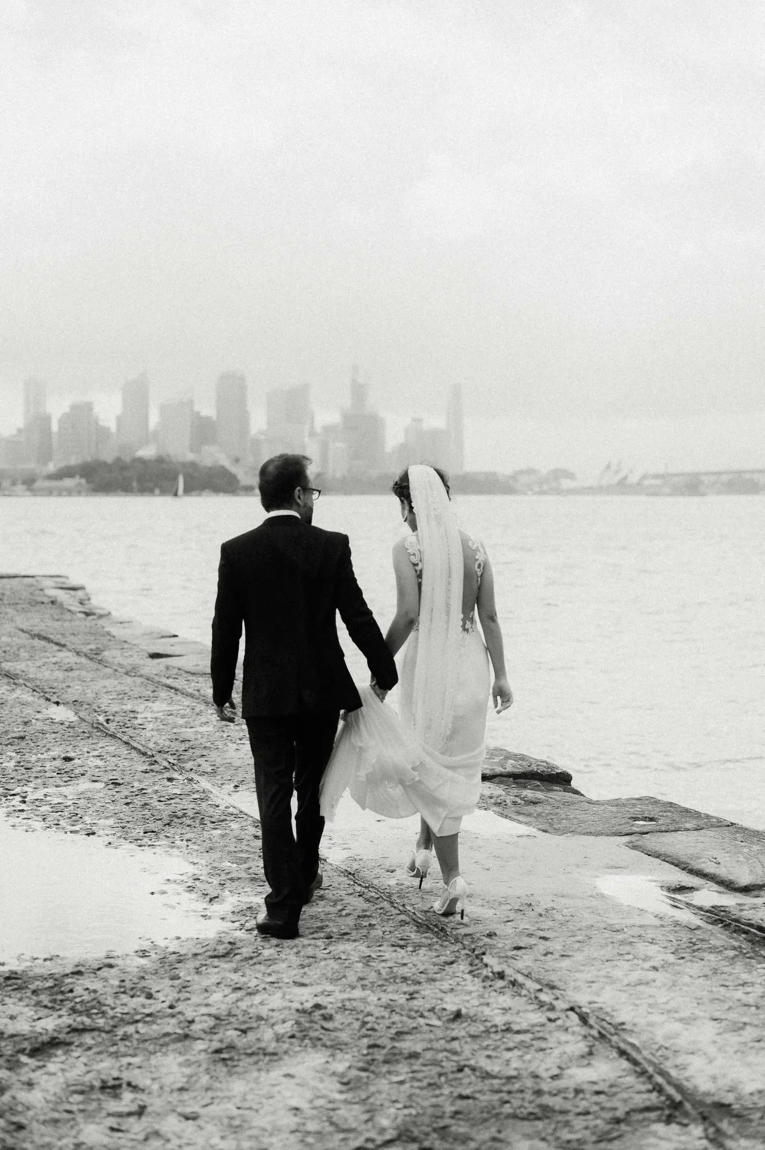 Black and white photograph of a bride and groom walking hand in hand along a waterfront promenade with a city skyline in the background.