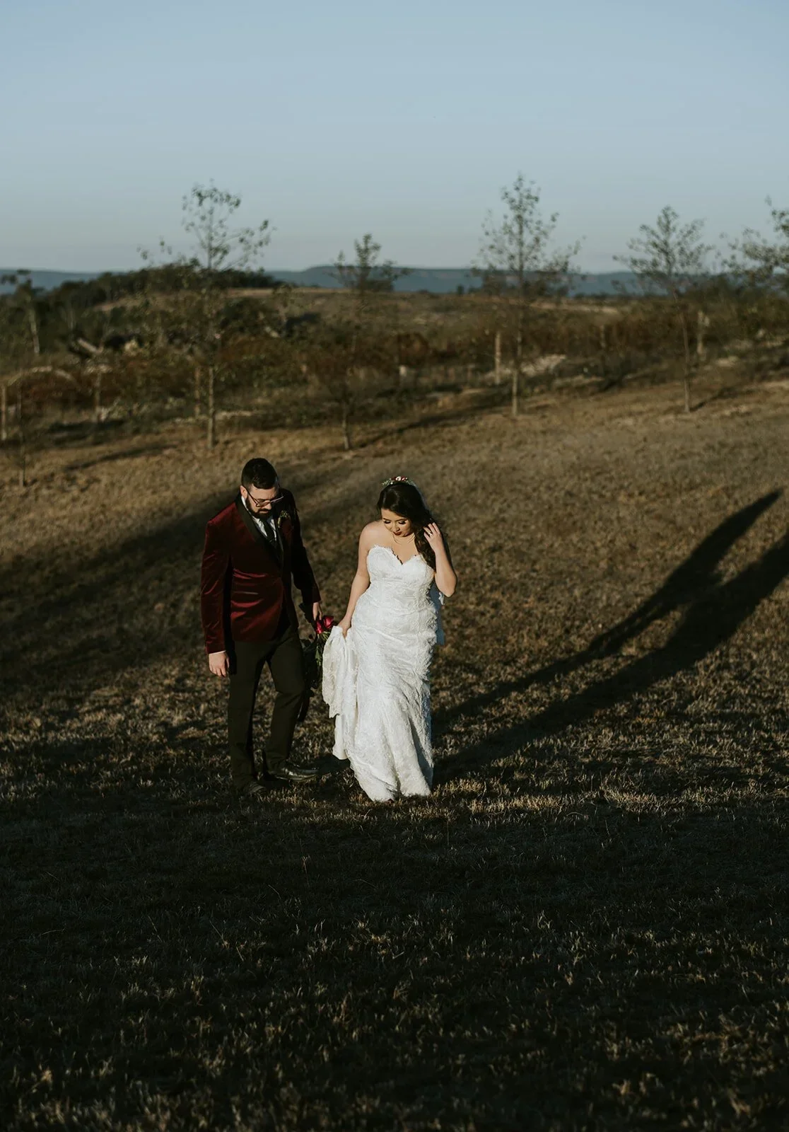 A bride in a white wedding dress and a groom in a dark suit holding hands and walking in an open field during sunset.