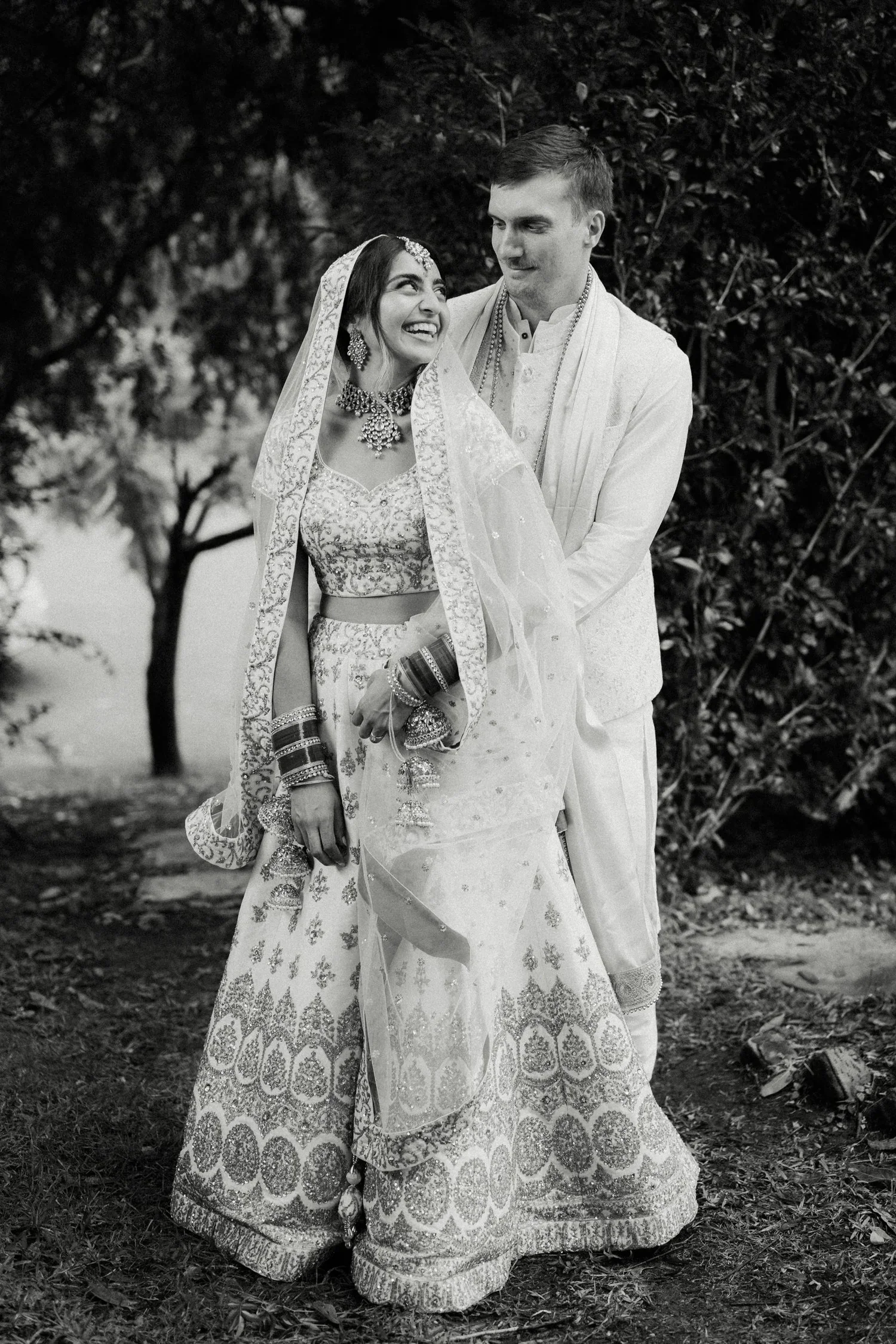 A bride and groom dressed in traditional Indian wedding attire outdoors, smiling at each other, with trees in the background.