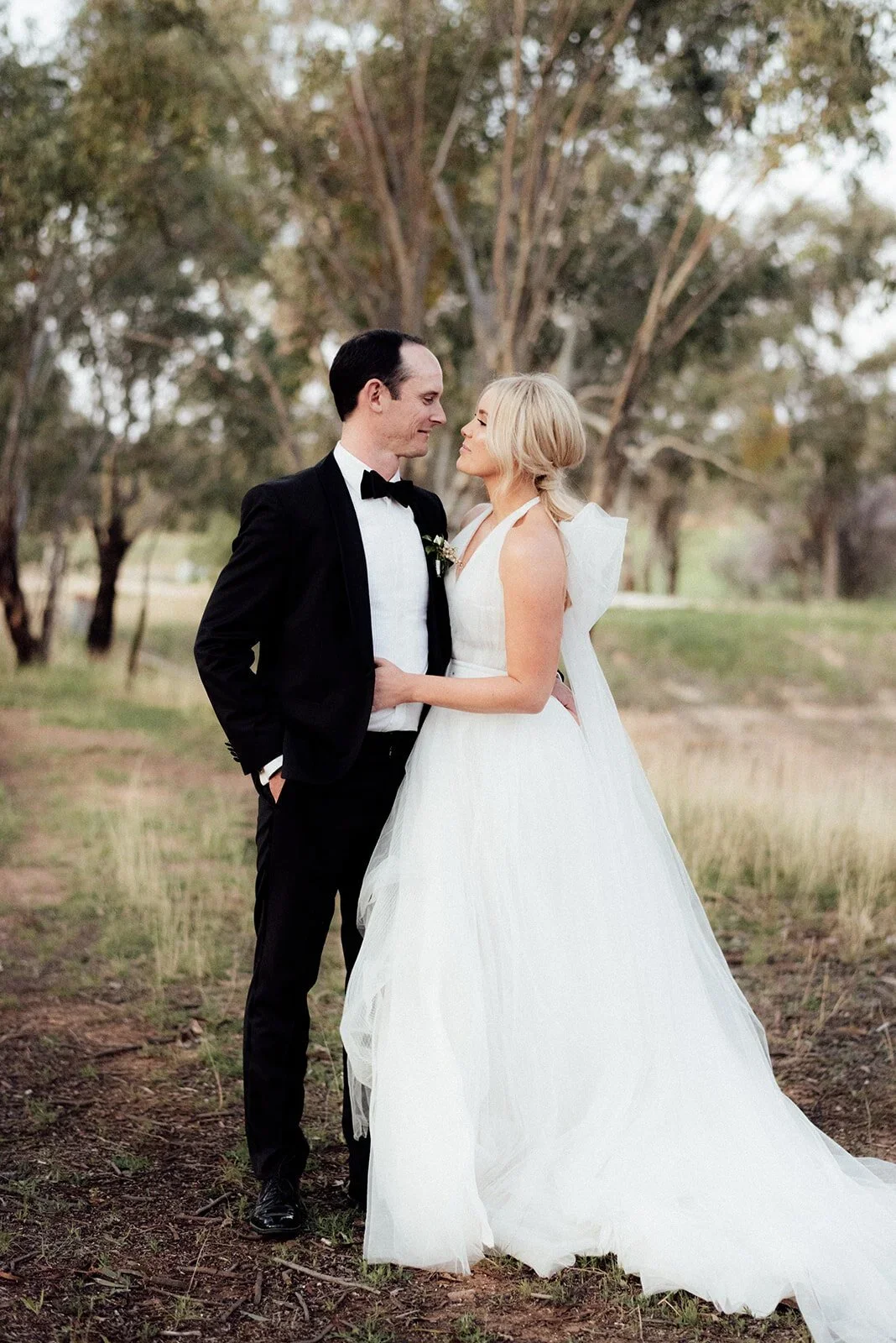 A newlywed couple stands close in an outdoor setting with trees in the background. The groom is wearing a black tuxedo with a bow tie and the bride is in a white wedding gown with ruffles on the shoulders.