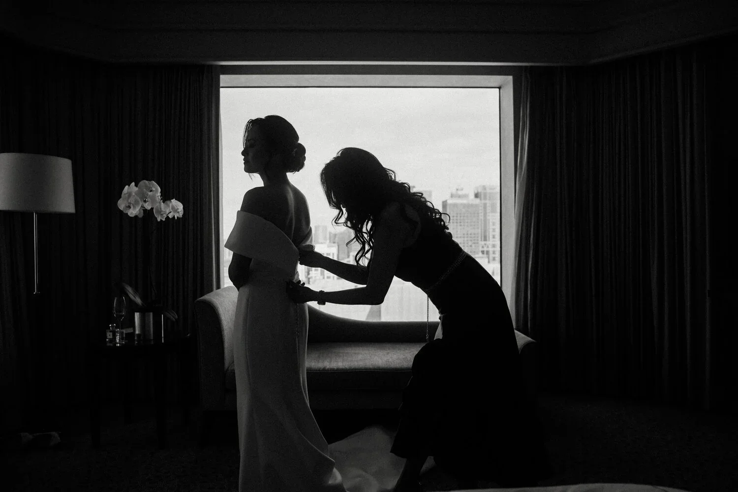 Silhouette of two women in a hotel room, one adjusting the other's dress, with a city view through a large window in the background.