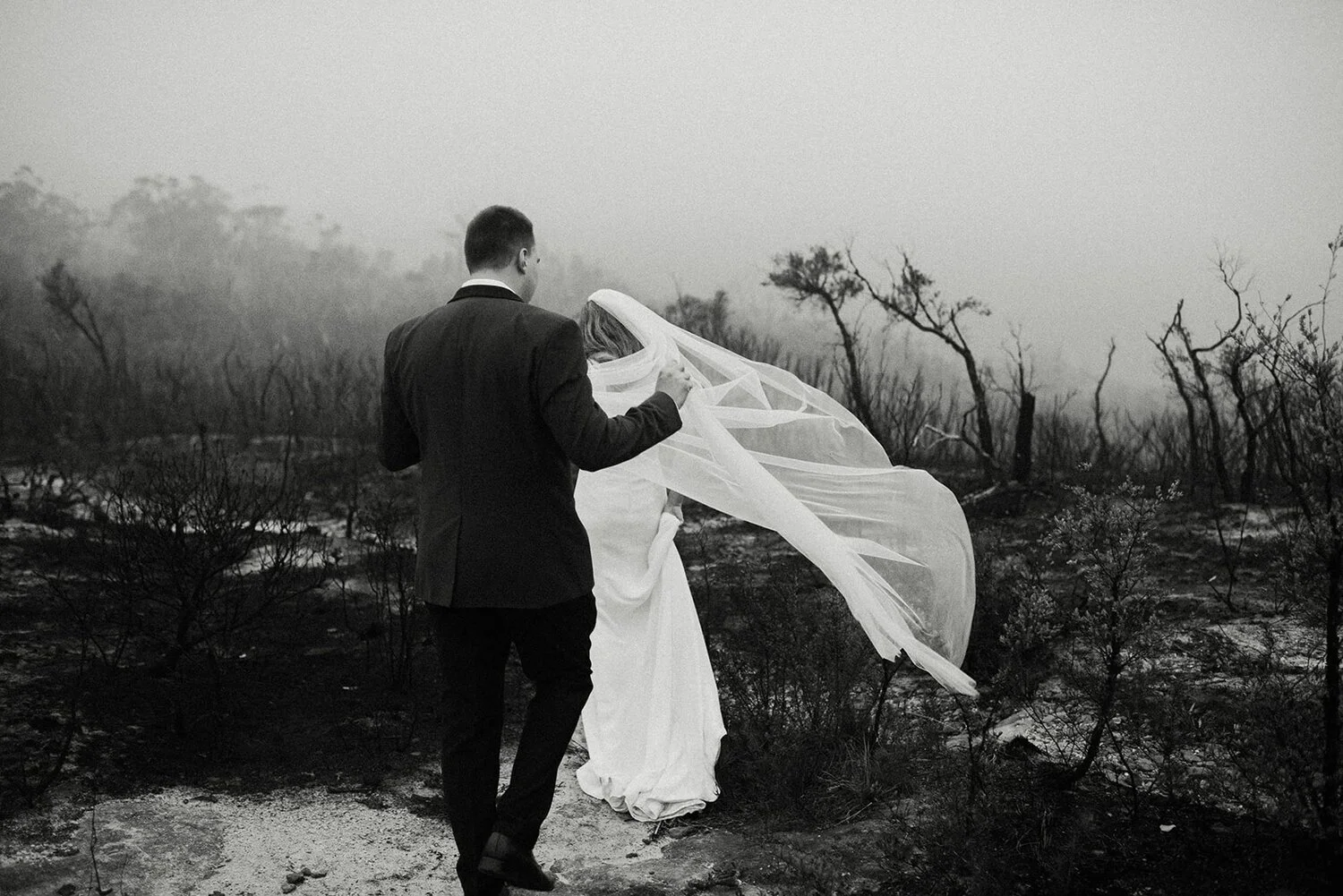 A black and white photo of a bride and groom in a smoky, desolate landscape. The groom stands behind the bride, holding her veil, as they walk away from the camera.