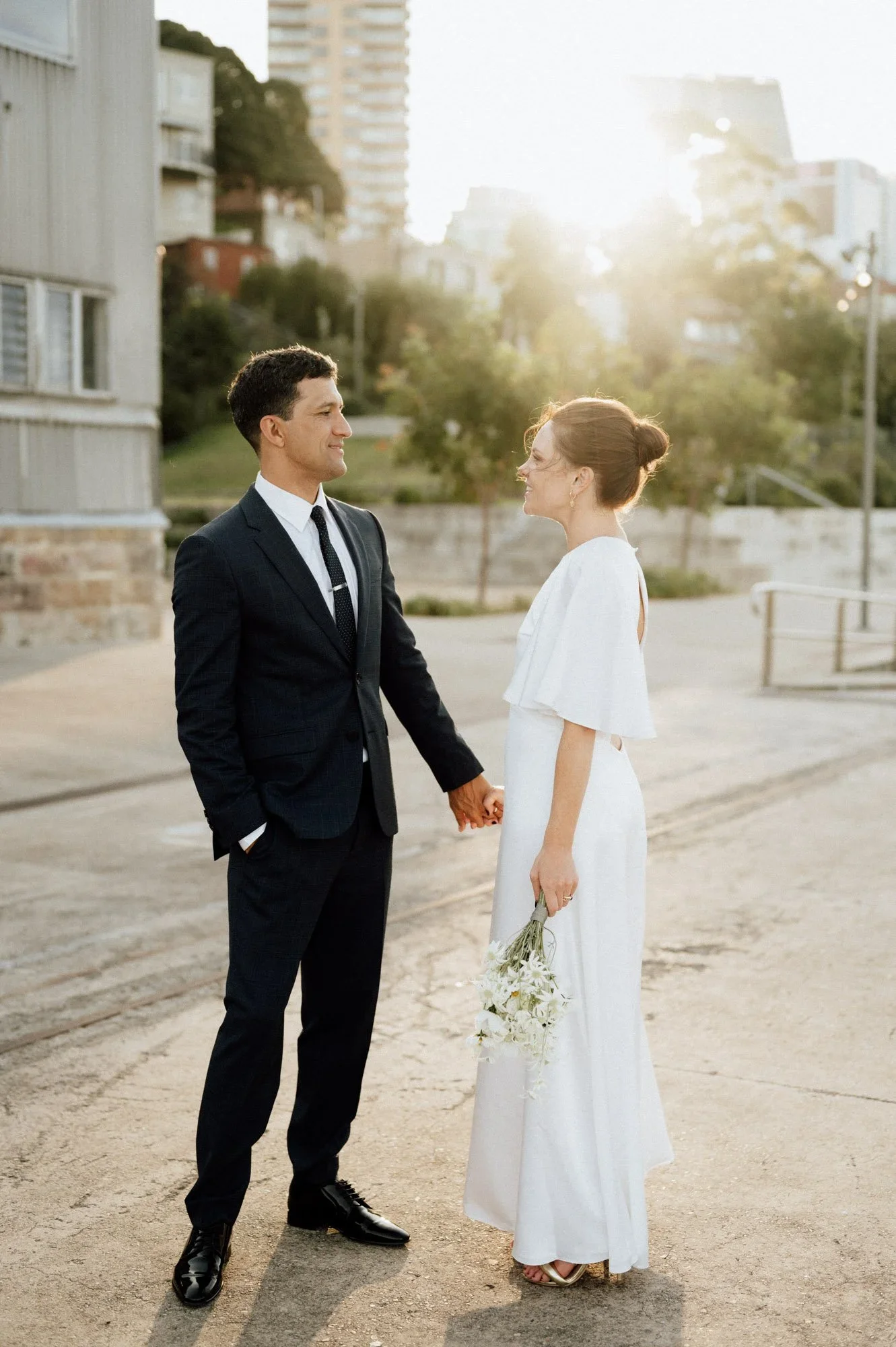 A man and woman dressed in wedding attire holding hands and smiling at each other outdoors, with city buildings in the background and the sun shining brightly.