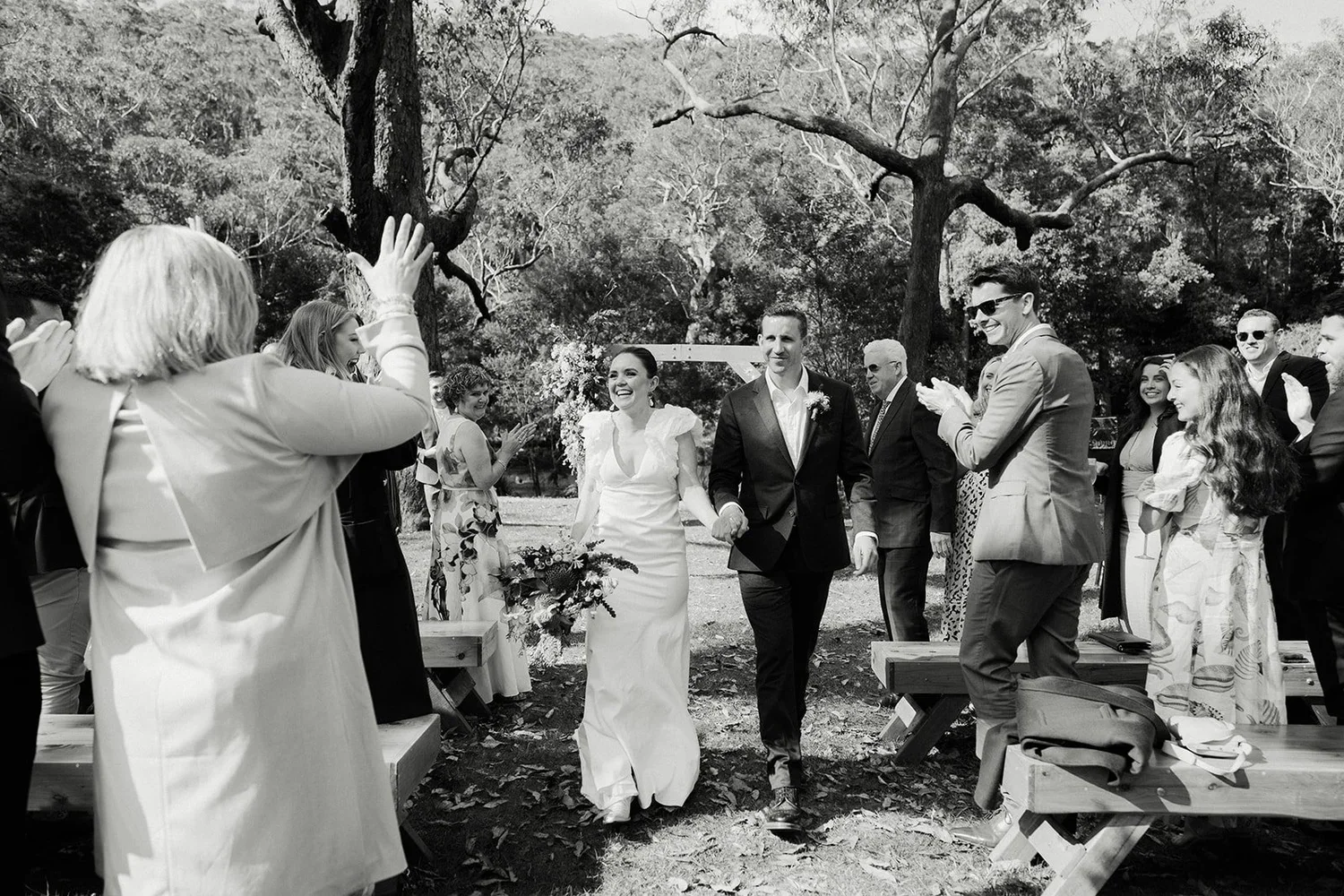 Black and white photo of a bride and groom walking down the aisle hand in hand at an outdoor wedding ceremony, surrounded by guests clapping and celebrating.