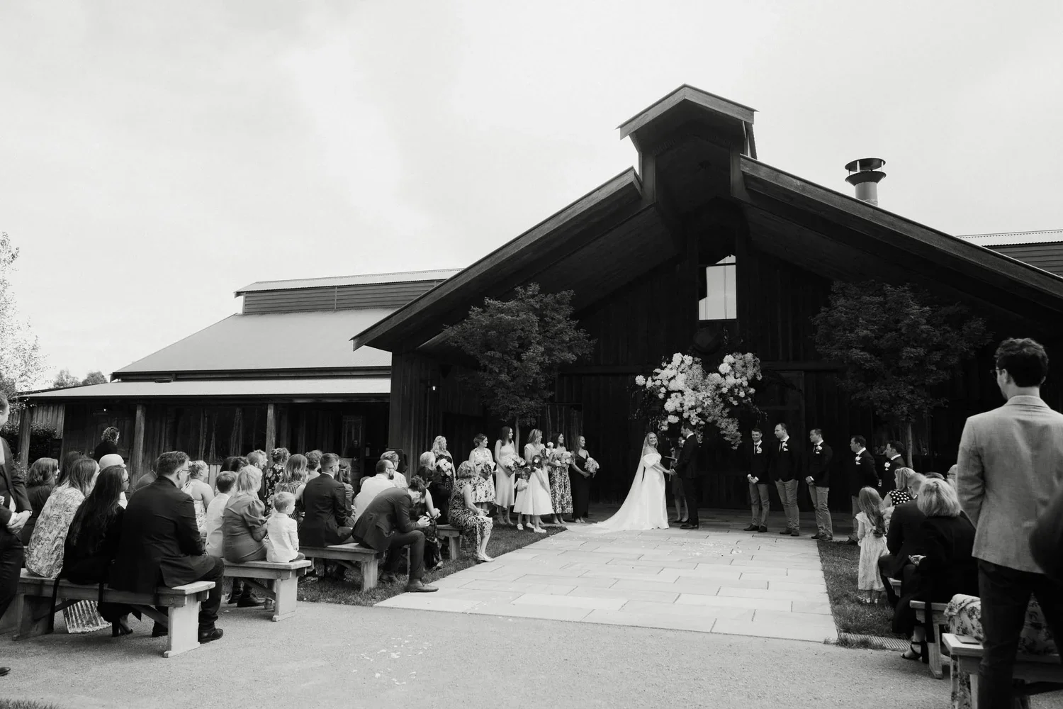 Couple getting married during outdoor wedding ceremony in front of guests seated on benches in front of a rustic black barn, decorated with flowers and trees.