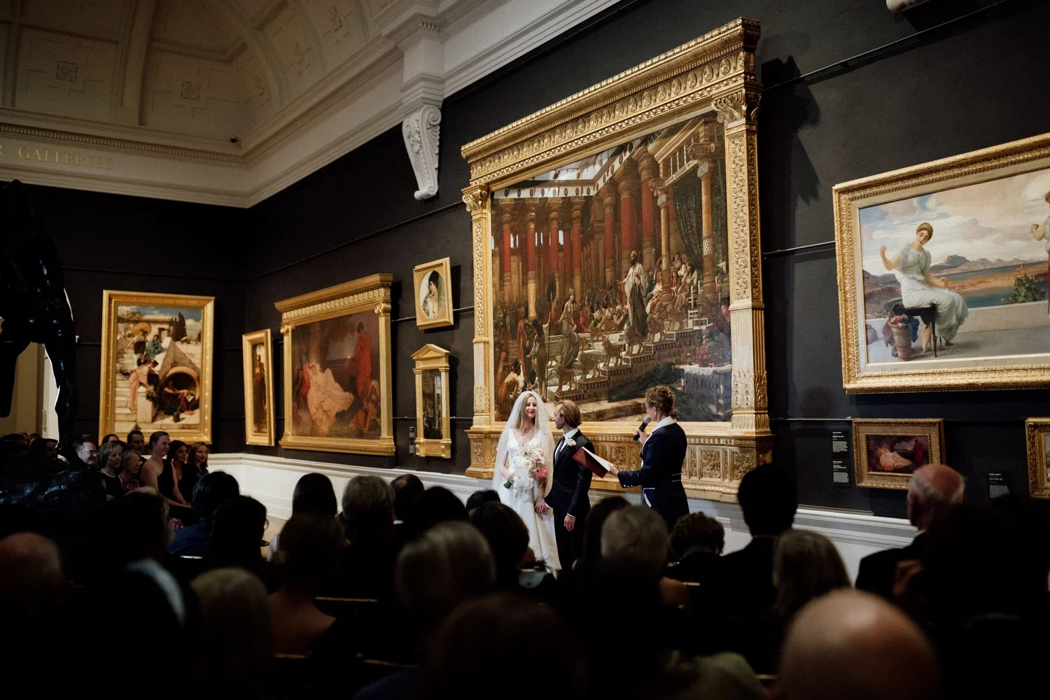 A wedding ceremony taking place in an art gallery with paintings on the wall. The bride and groom stand facing each other, while a person officiates with a microphone and a book. The audience is seated and watching the ceremony.