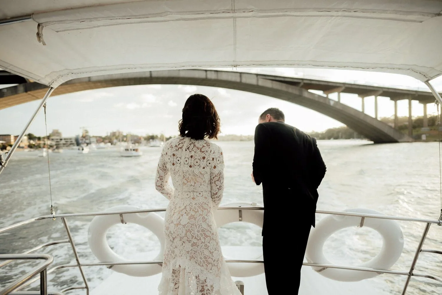 A woman in a lace dress and a man in a suit on a boat, looking out over the water near a bridge and boats in the distance during sunset.