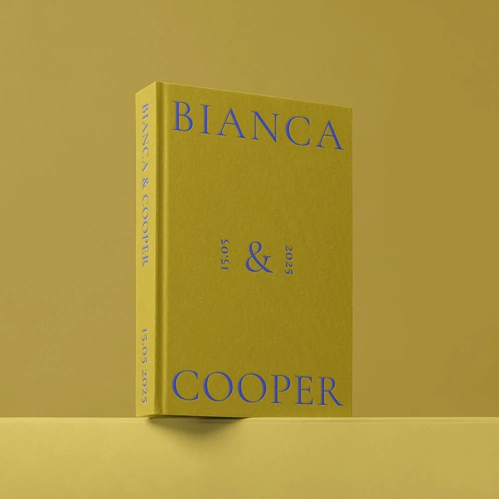 Yellow book with blue text titled 'Bianca & Cooper' standing upright on a yellow surface against a yellow background, with event date and time printed on the cover and spine.