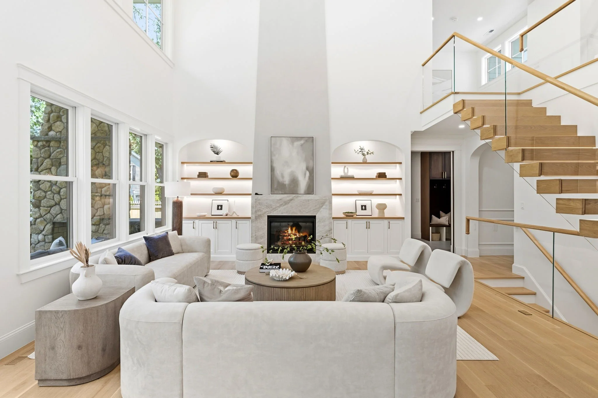 A bright, modern living room with large windows, a fireplace, and a staircase with wooden steps and glass railing.