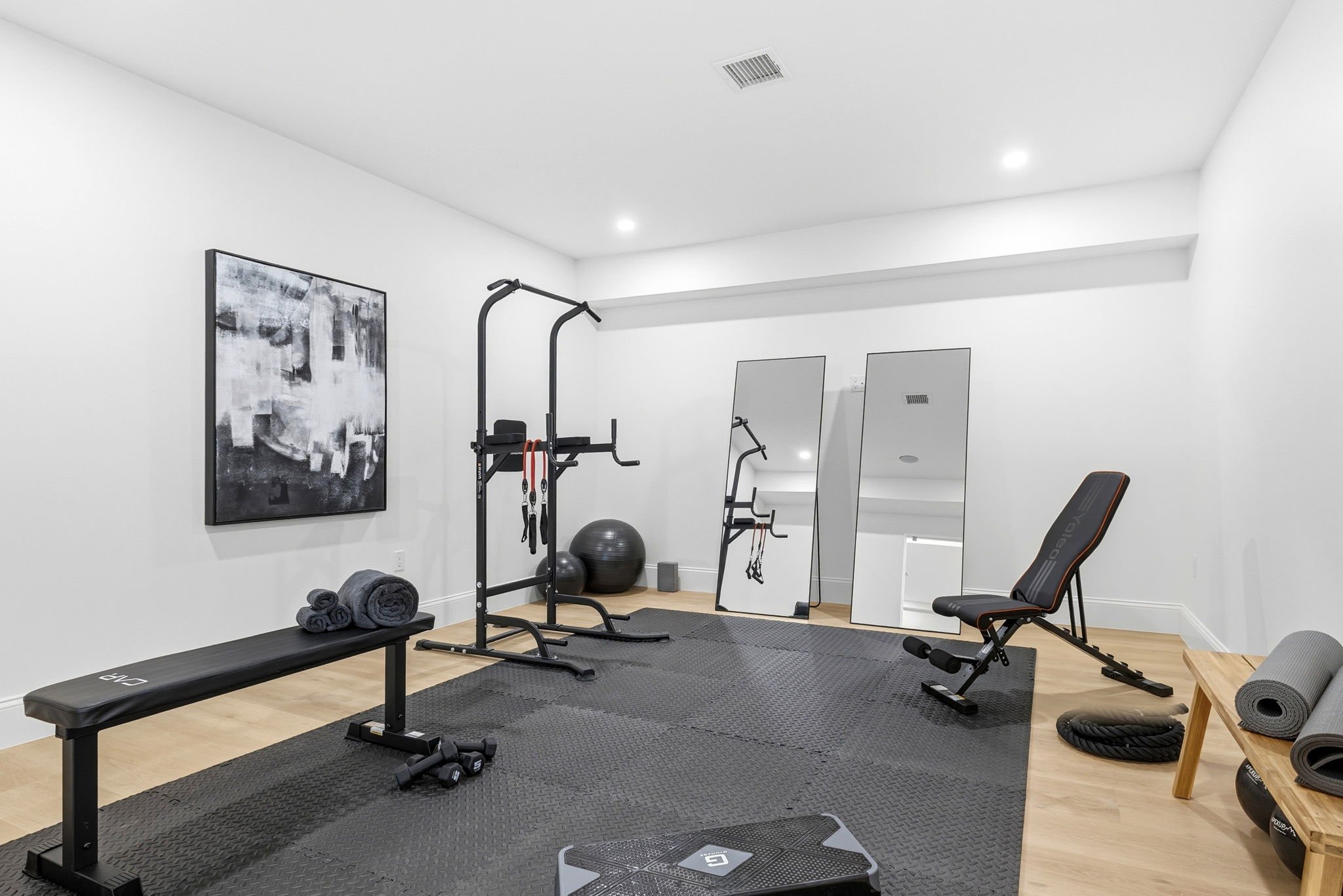 A home gym with white walls and ceiling, featuring exercise equipment including a black weight bench, dumbbells, stability balls, pull-up bar, yoga mats, and a reclining exercise chair. There are framed abstract artwork and mirrors on the walls.