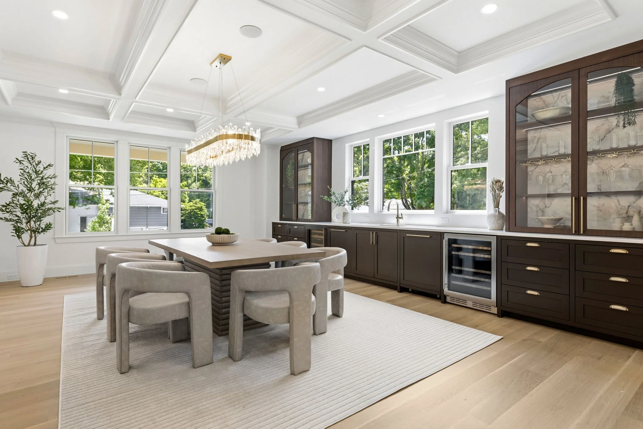Bright, modern kitchen and dining area with large windows, dark cabinetry, a marble-topped cabinet, and a chandelier over the dining table.