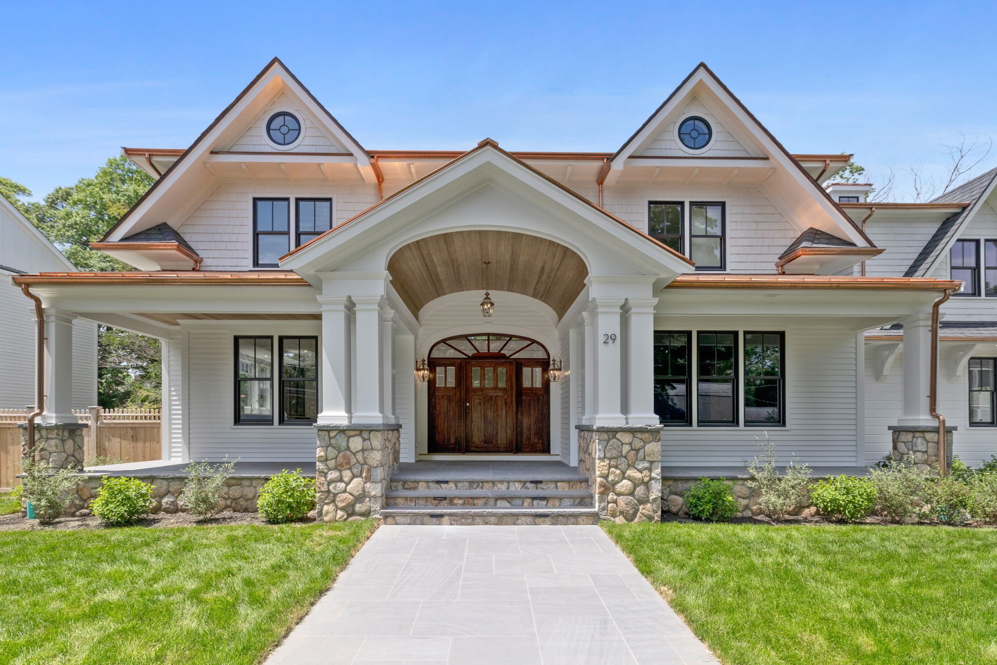Front view of a modern white house with stone accents, a covered porch, and a pathway leading to the entrance, surrounded by green grass and plants.