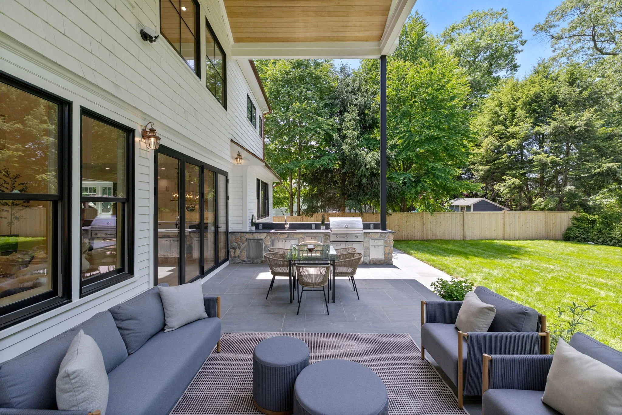 A modern outdoor patio with gray cushioned chairs and a dining table, adjacent to a house with sliding glass doors, a built-in barbecue, and surrounded by green trees and grass.