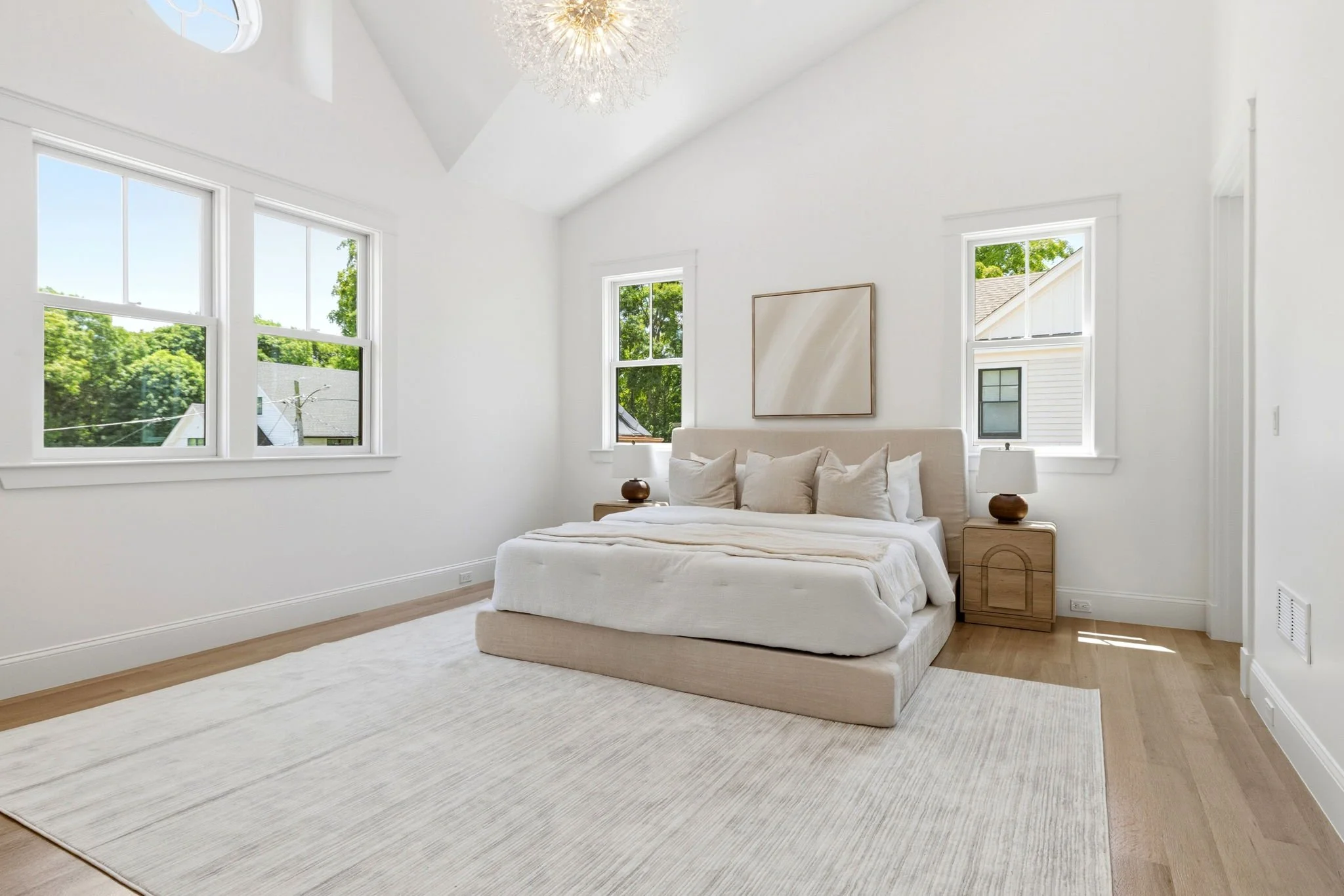 A minimalist bedroom with a large cream-colored bed, two matching nightstands with white lamps, three pillows, a blank wall painting, next to four large windows showing green trees outside, white walls, and light wood flooring.