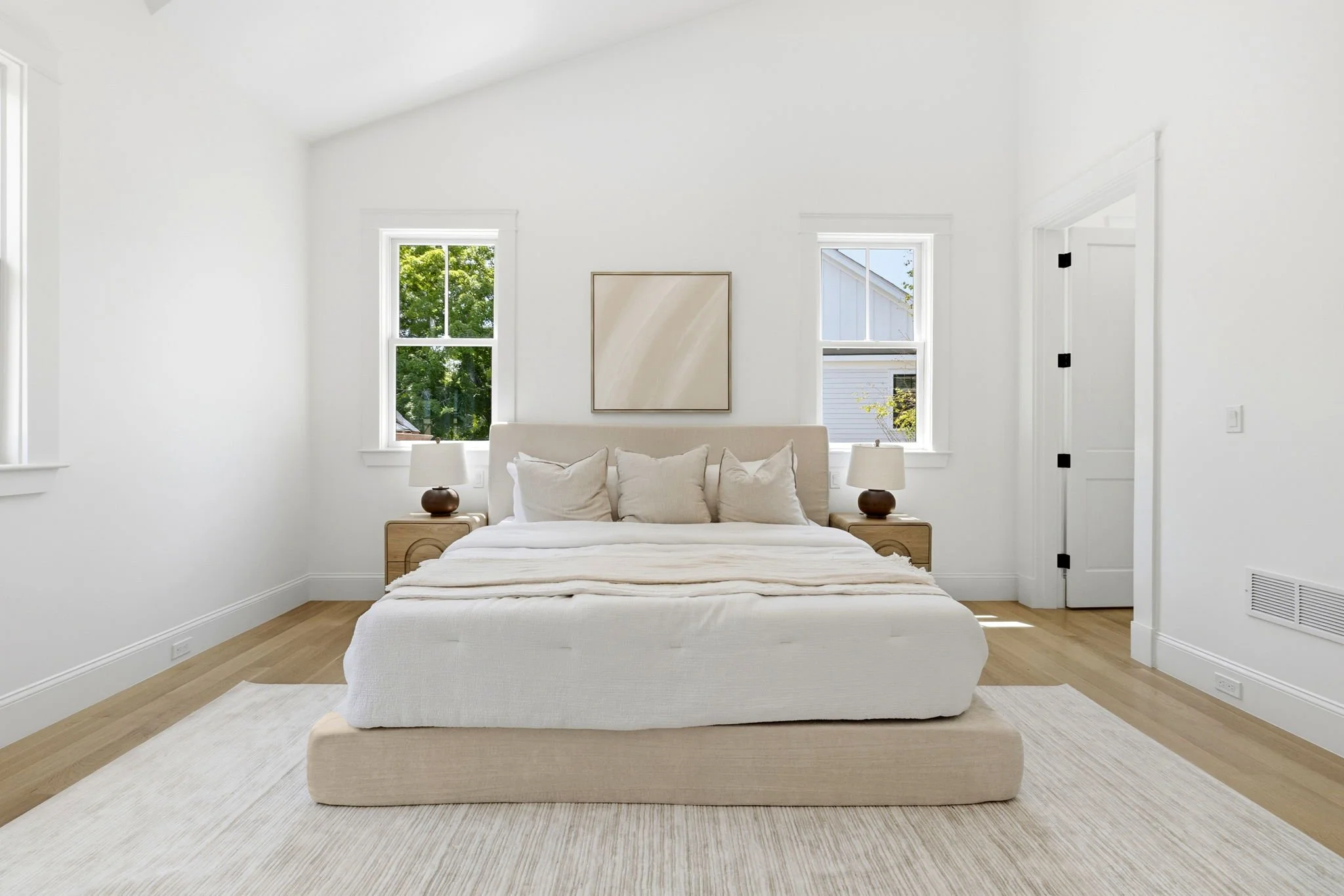 A minimalist bedroom with white walls, a beige upholstered bed with white pillows, two matching bedside tables with lamps, two windows with greenery outside, and simple artwork on the wall.